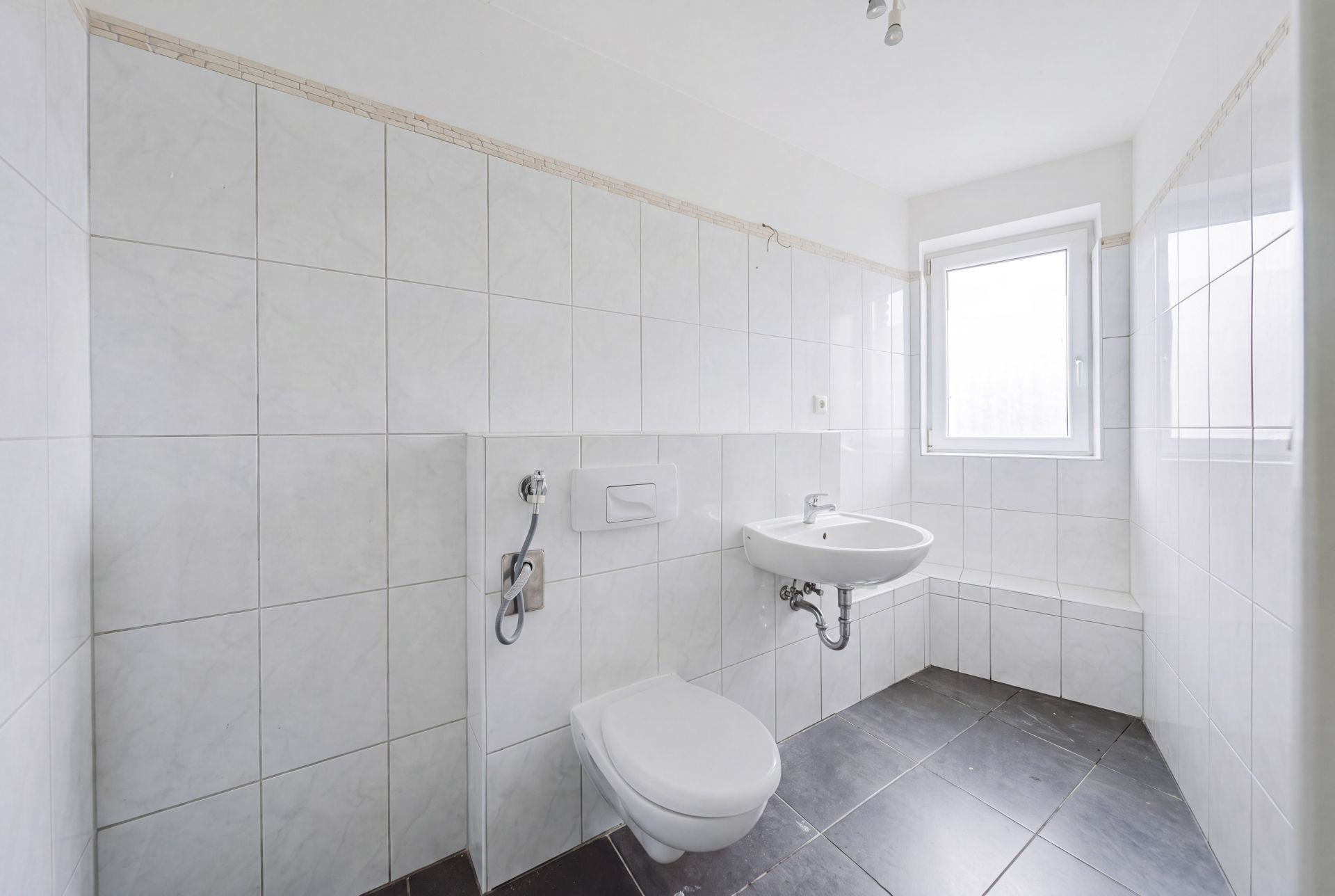 Helles, gefliestes Badezimmer mit Wandtoilette, Waschbecken und Fenster.
