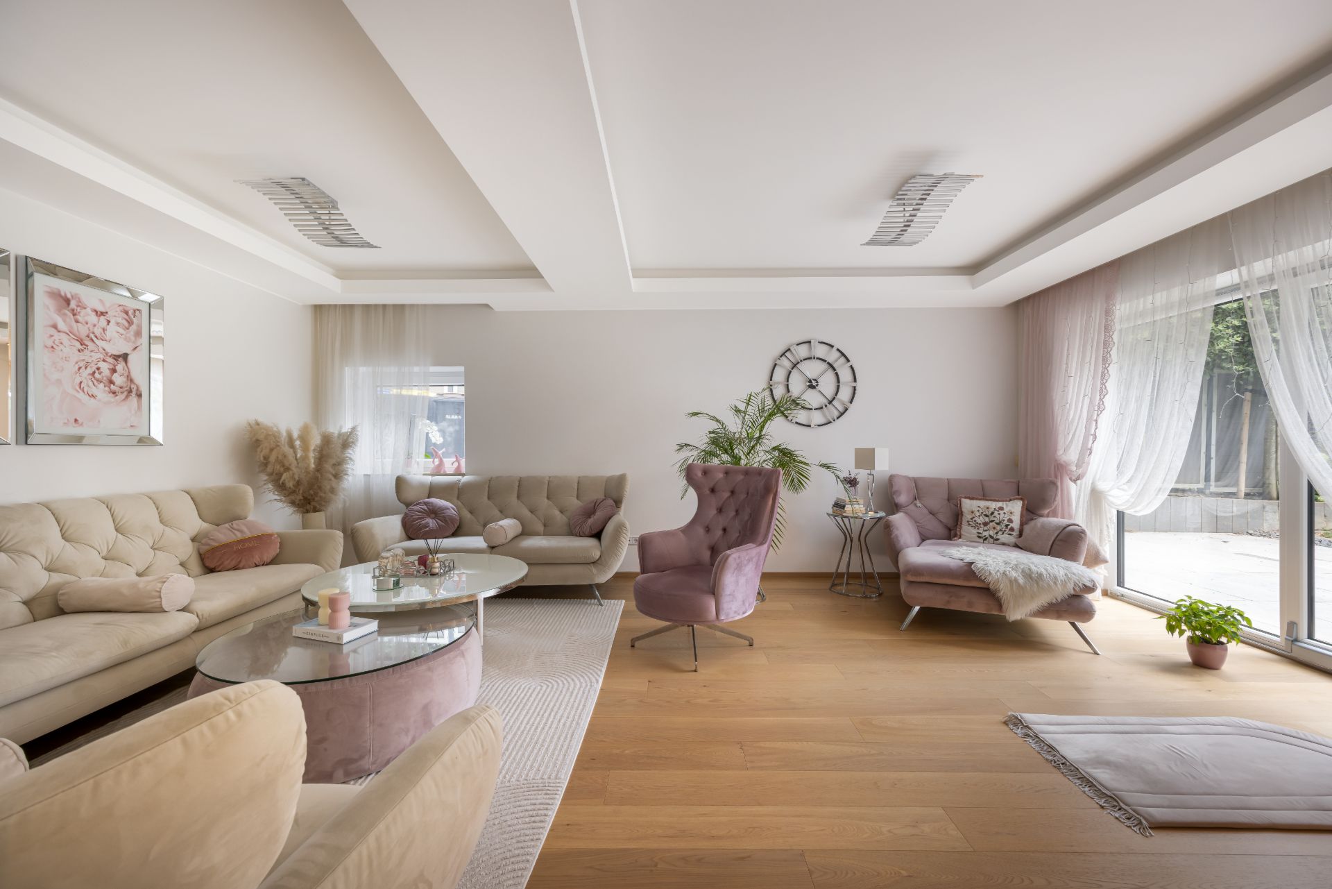 Helles Wohnzimmer mit beigen Sofas, Glastisch, lilafarbener Deko und großen Fenstern mit Gardinen.