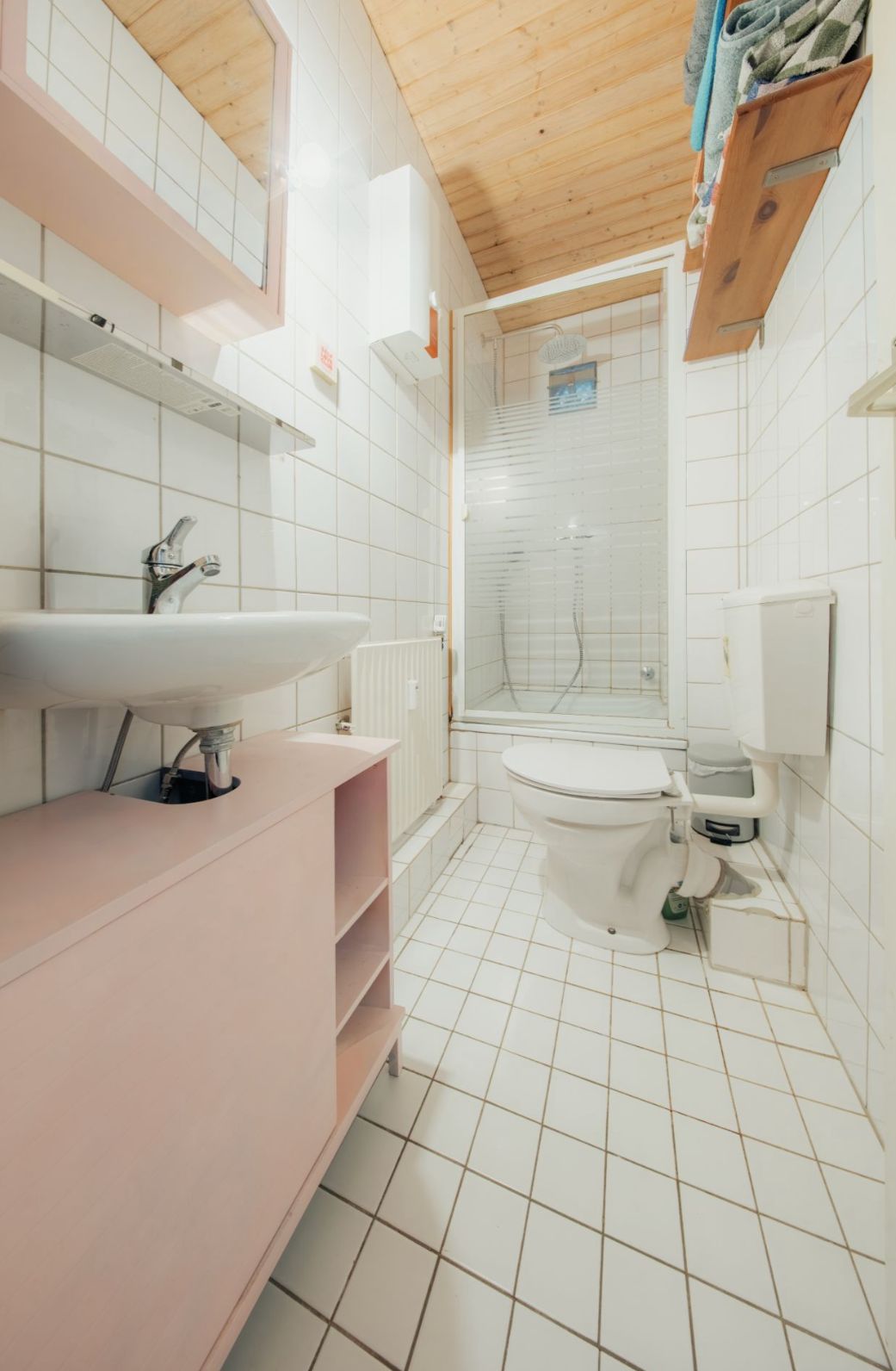 Helles Bad mit Waschbecken, Toilette, Dusche und Fliesenboden, Holzdecke oben.