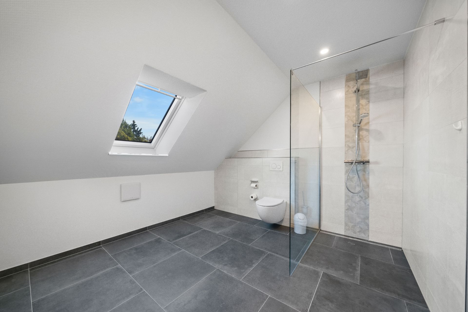 Modernes Bad mit Dachfenster, bodengleicher Dusche, WC und grauen Fliesen.