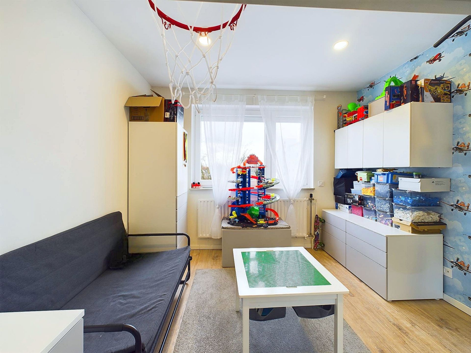 Helles Kinderzimmer mit Spielzeug, Sofa, Wandregalen und einem Basketballkorb an der Decke.