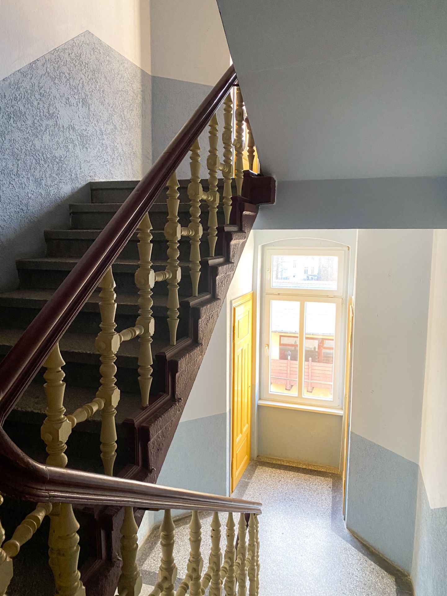 Treppenhaus mit Holzgeländer, Fenster und gelber Tür auf einer Etage.