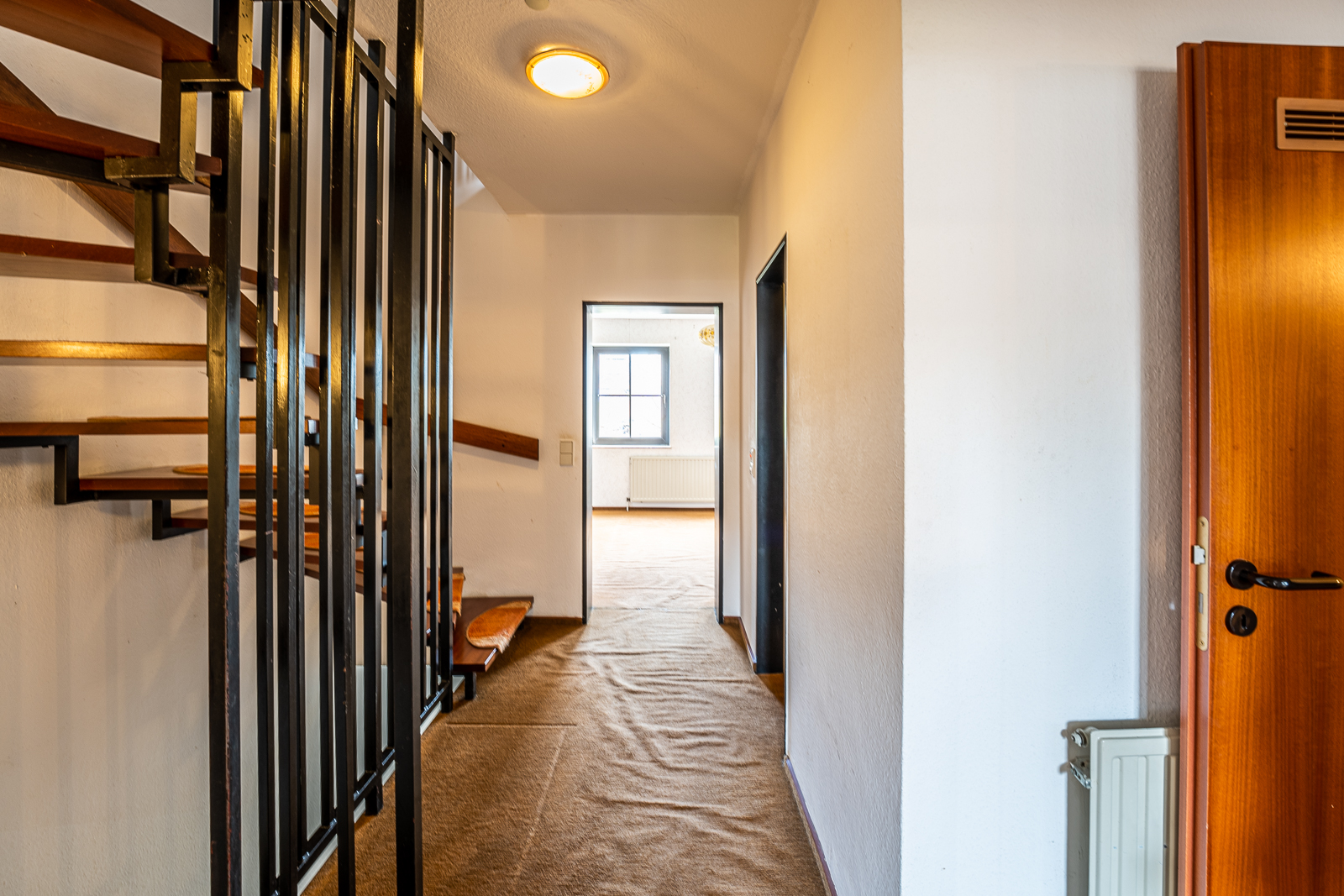 Flur mit Teppichboden, offener Holztreppe und Blick in ein helles Zimmer mit Fenster.