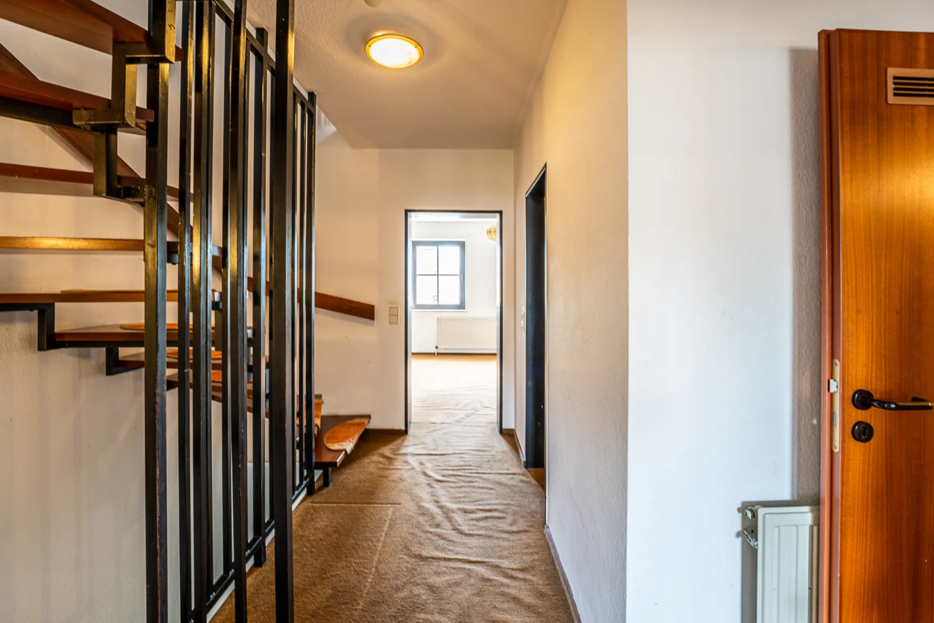 Flur mit Teppichboden, offener Holztreppe und Blick in ein helles Zimmer mit Fenster.