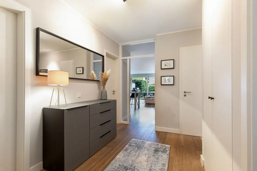 Heller Flur mit Kommode, Spiegel, Lampe, Teppich und Blick ins Wohnzimmer.