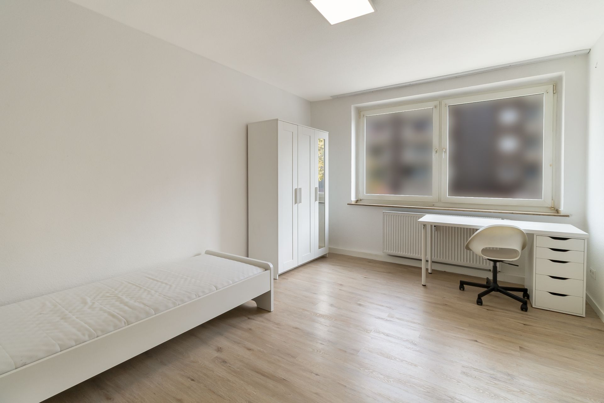 Helles Zimmer mit Einzelbett, Kleiderschrank, Schreibtisch und Stuhl, großes Fenster.