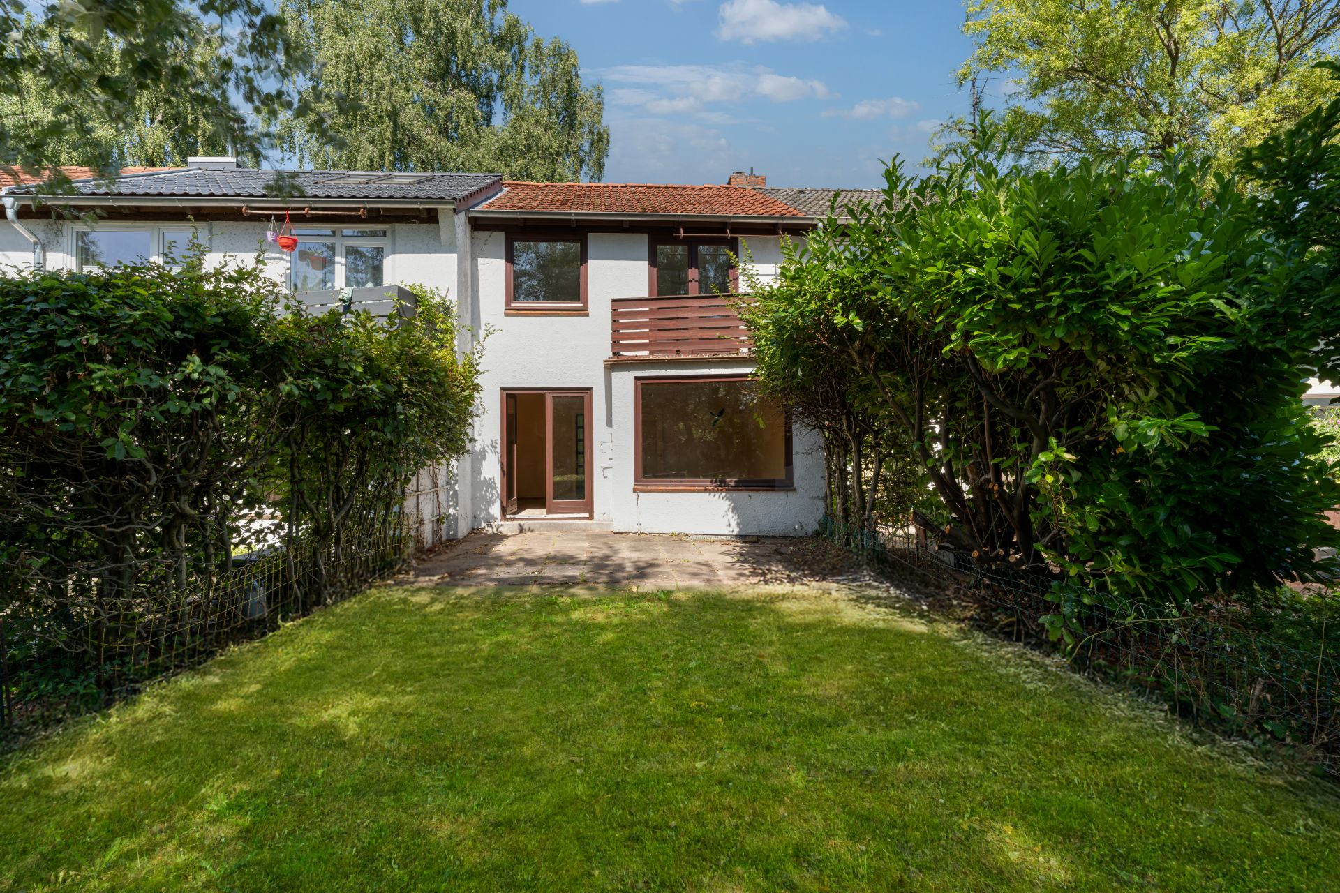 Zweistöckiges Haus mit Terrasse und Garten, umgeben von Büschen und Bäumen.