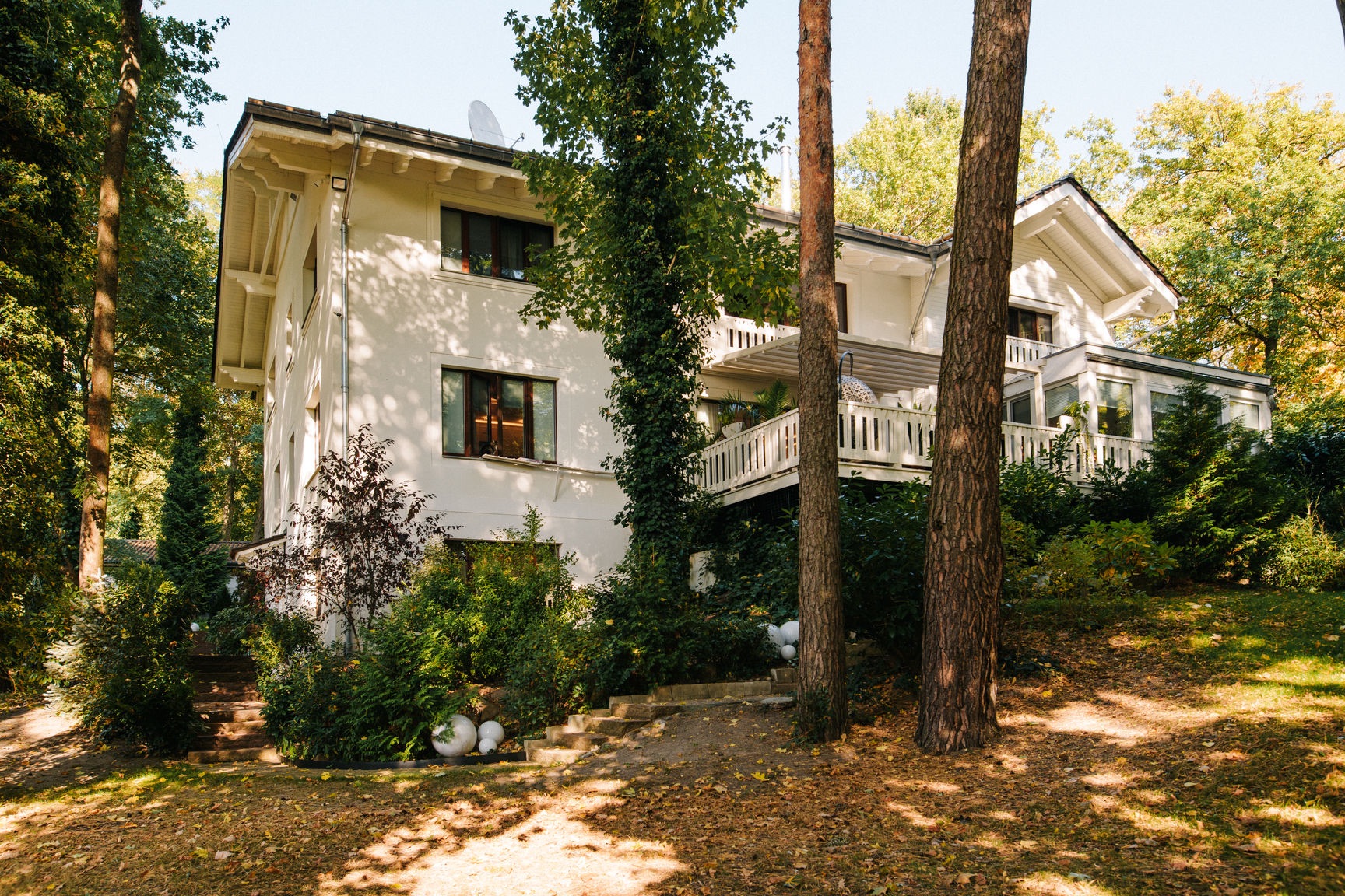 Weißes Wohnhaus mit Balkon, umgeben von Bäumen und Grünpflanzen im Garten.