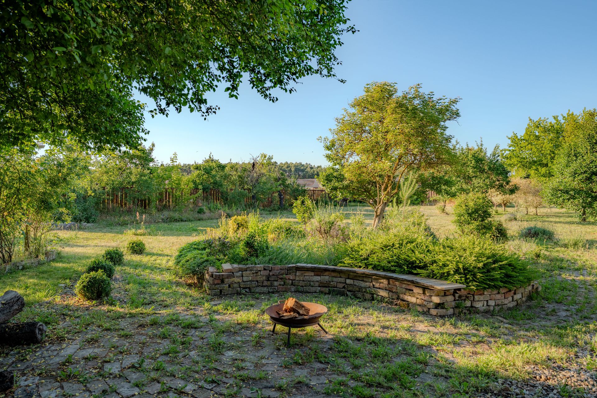 Garten mit Feuerstelle, gebogener Steinmauer und vielen Bäumen im Hintergrund.