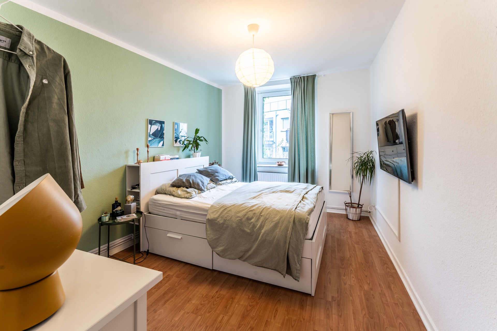 Helles Schlafzimmer mit Doppelbett, Holzfußboden, Wandspiegel, TV und Fenster mit Vorhängen.
