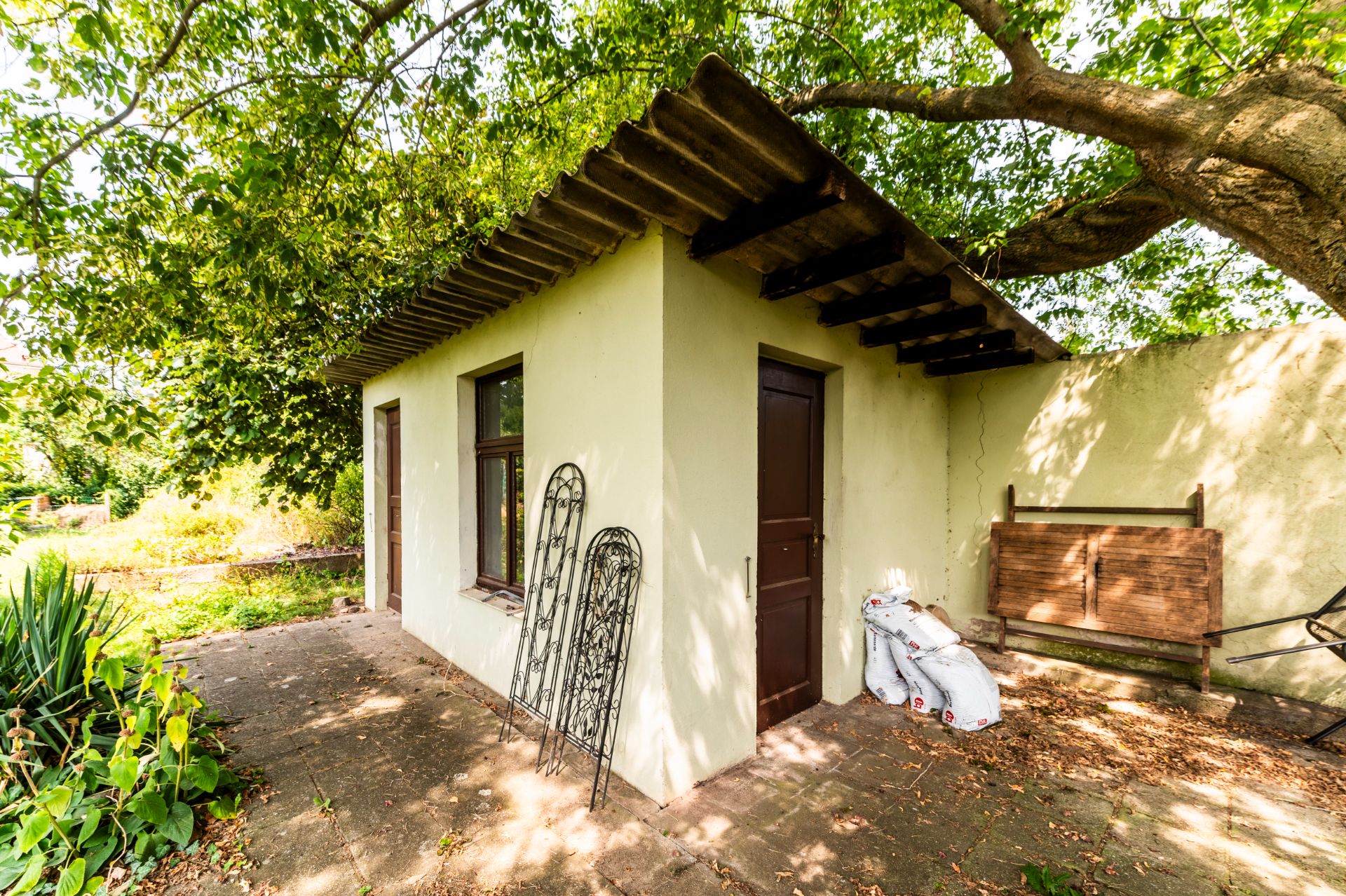 Kleines Gartenhaus mit Tür und Fenster, umgeben von Bäumen und Pflanzen.