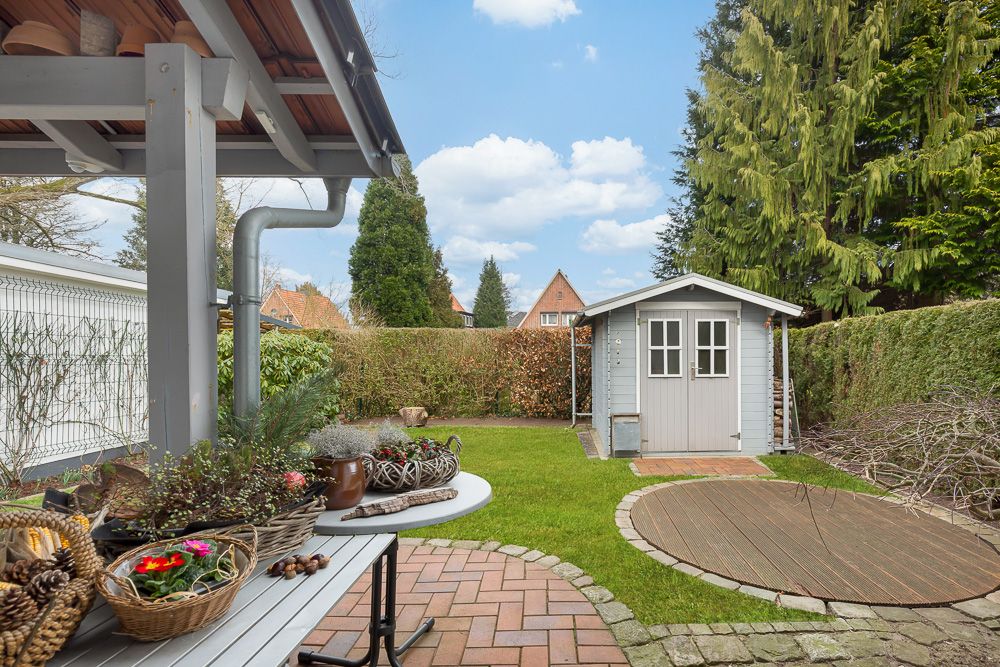 Garten mit Rasen, Holzhütte, Steinterrasse und Pflanzen auf einem Tisch im Vordergrund.