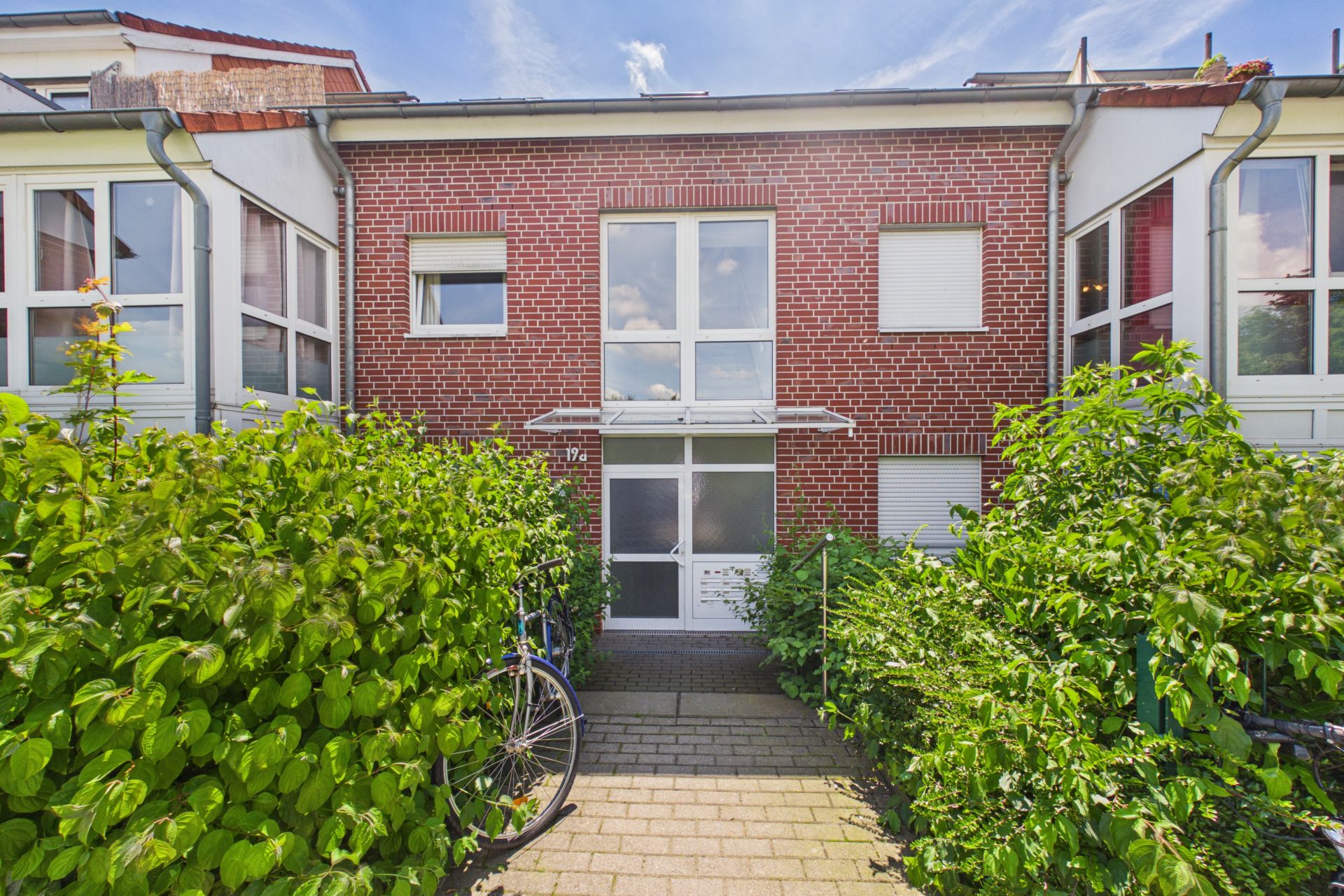 Eingang eines zweistöckigen Backsteinhauses mit weißen Fenstern, umgeben von grünen Büschen und einem Fahrrad.
