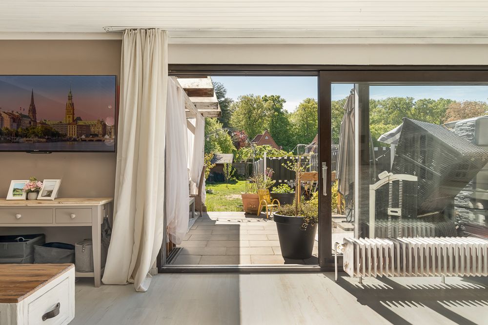 Wohnzimmer mit offener Terrassentür, Blick auf Terrasse und Garten mit Pflanzen.