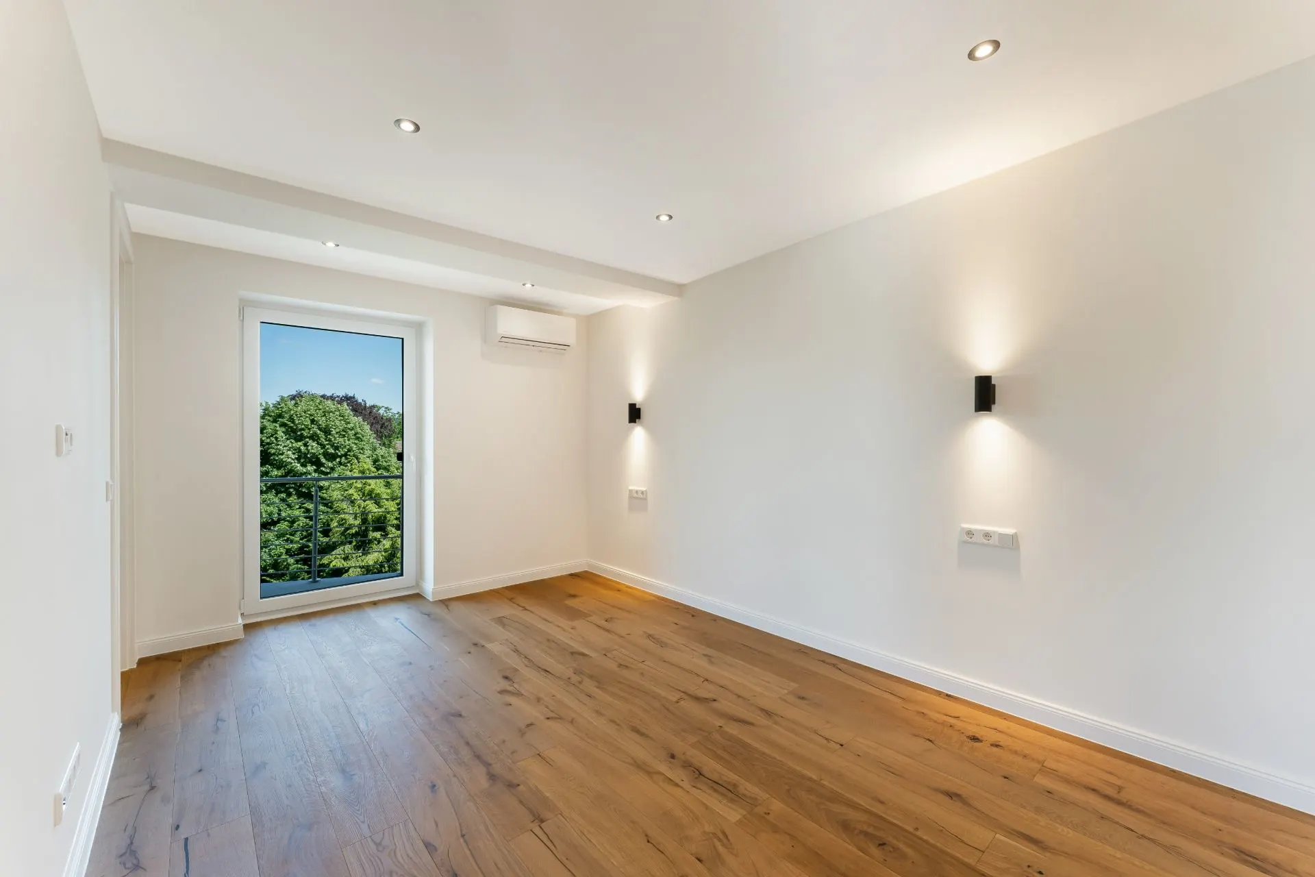 Helles, leeres Zimmer mit Holzboden, weißen Wänden und Fenster mit Blick ins Grüne.