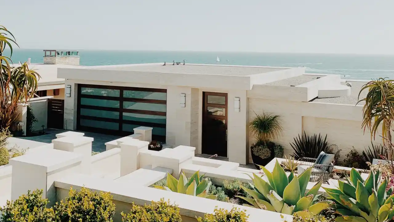 Modernes Haus mit Flachdach, großem Fenster und Pflanzen im Vorgarten, im Hintergrund das Meer.