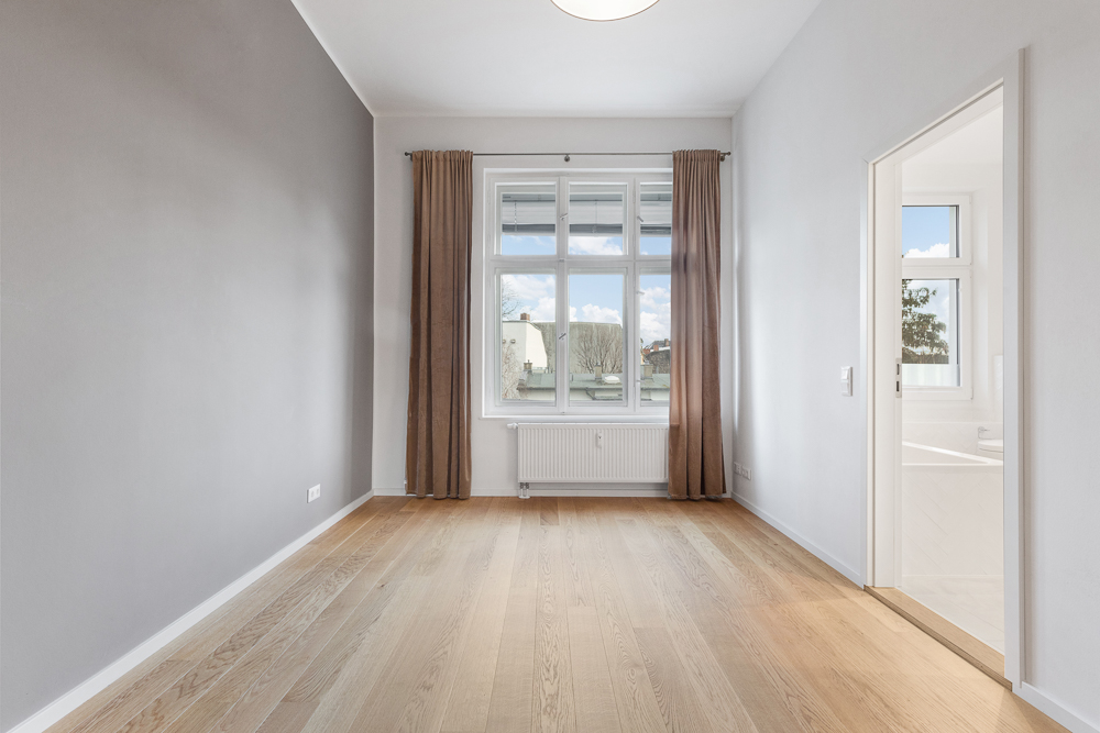 Leerer Raum mit Holzfußboden, großem Fenster und braunen Vorhängen, Tür führt in ein weiteres Zimmer.