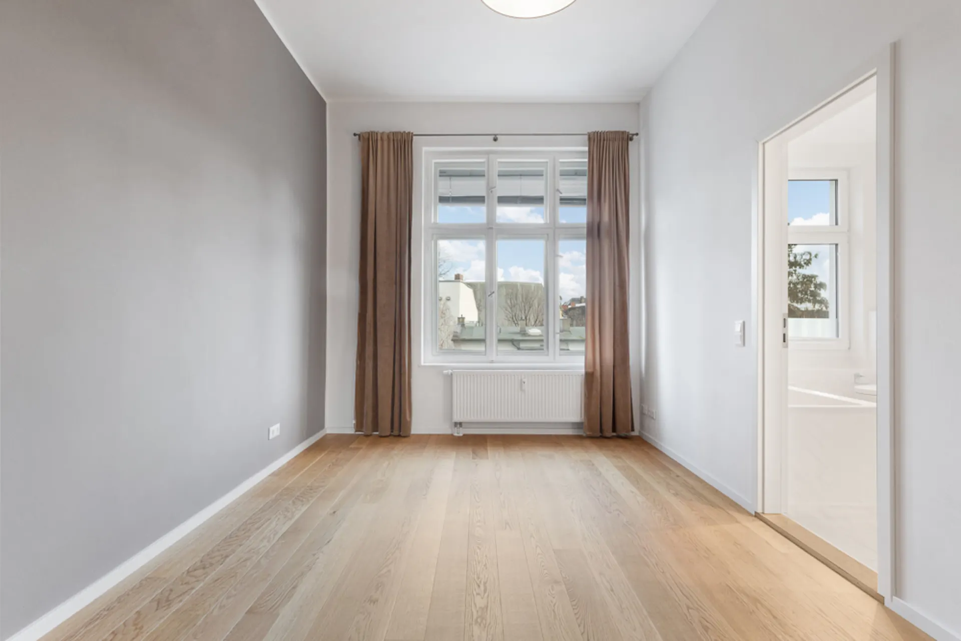 Leerer Raum mit Holzfußboden, großem Fenster und braunen Vorhängen, Tür führt in ein weiteres Zimmer.