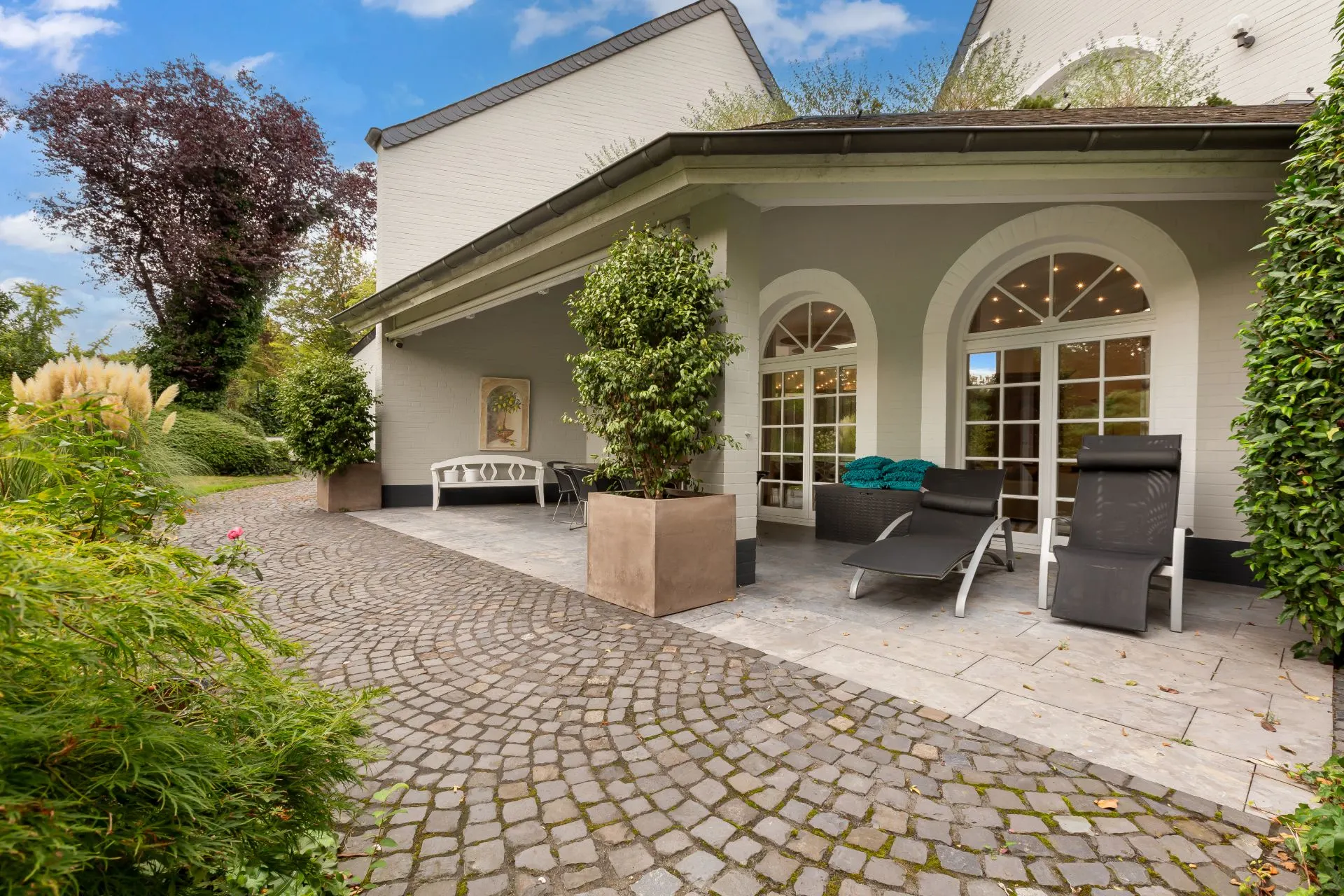 Gepflasterte Terrasse mit Liegestühlen, Tisch, Pflanzenkübeln und großen Fenstern an einem Wohnhaus.