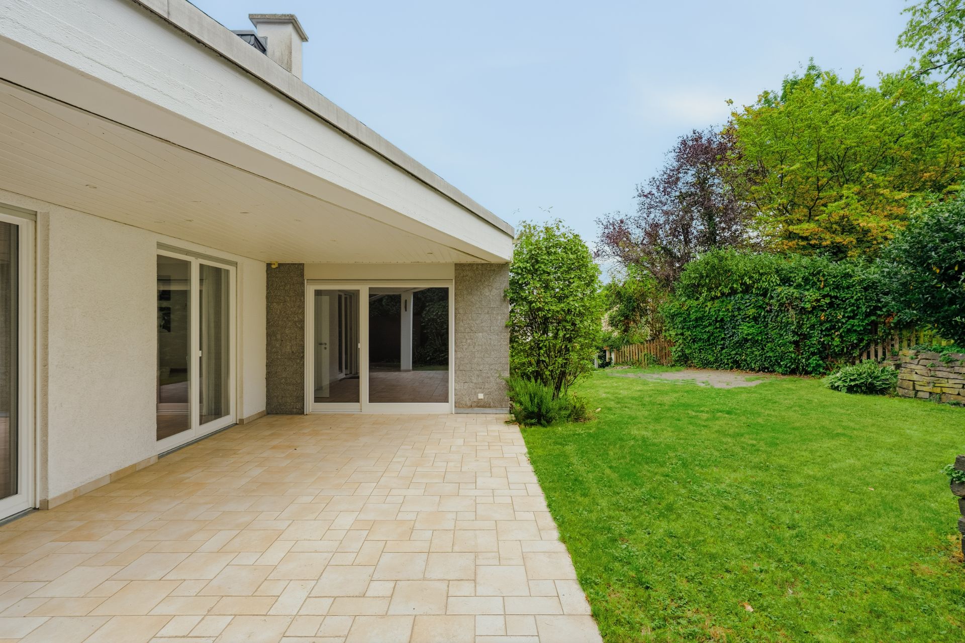 Überdachte Terrasse mit Fliesenboden und angrenzendem Garten, umgeben von Bäumen und Hecken.