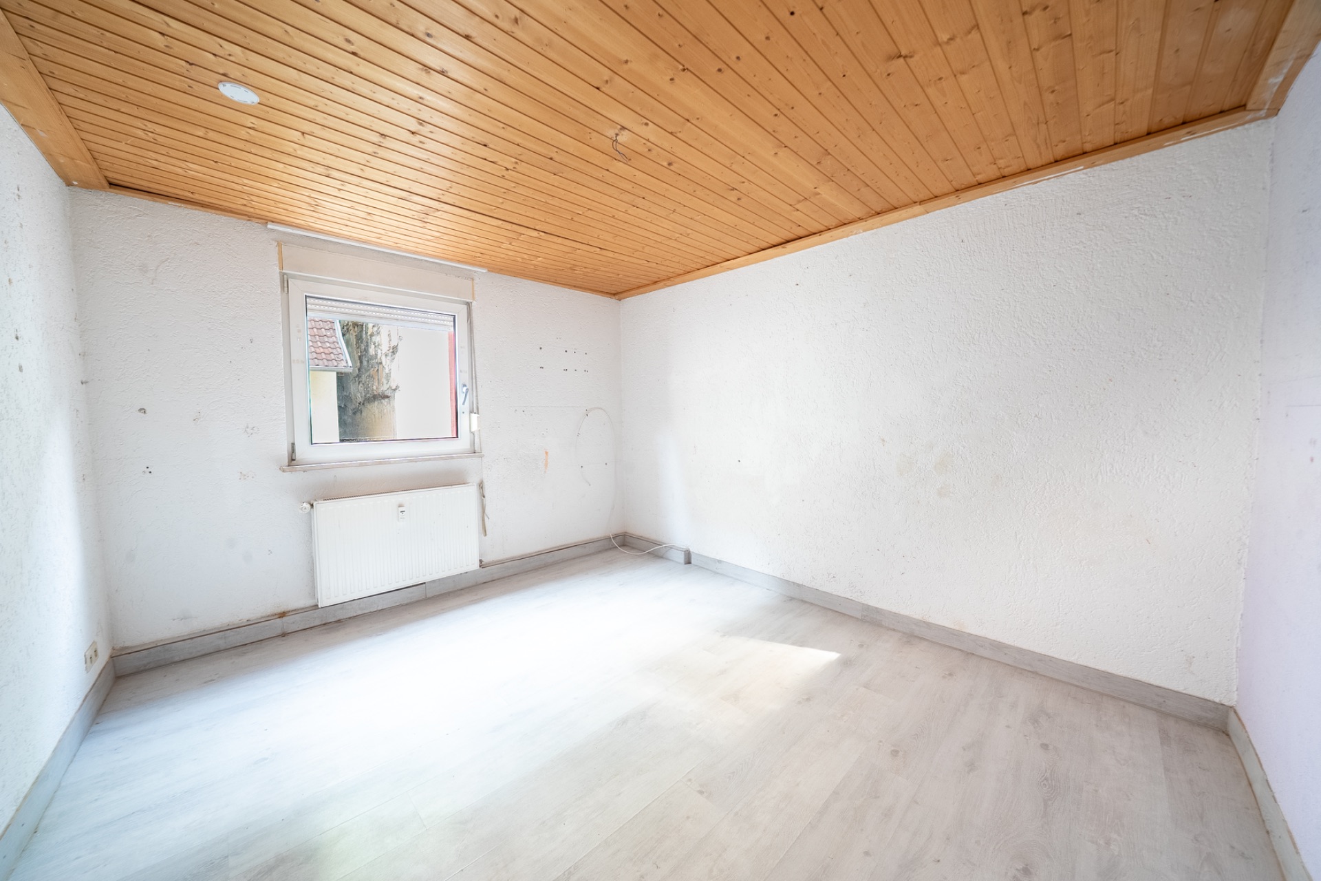 Leeres Zimmer mit weißem Boden, Holzdecke und Fenster, durch das Tageslicht einfällt.