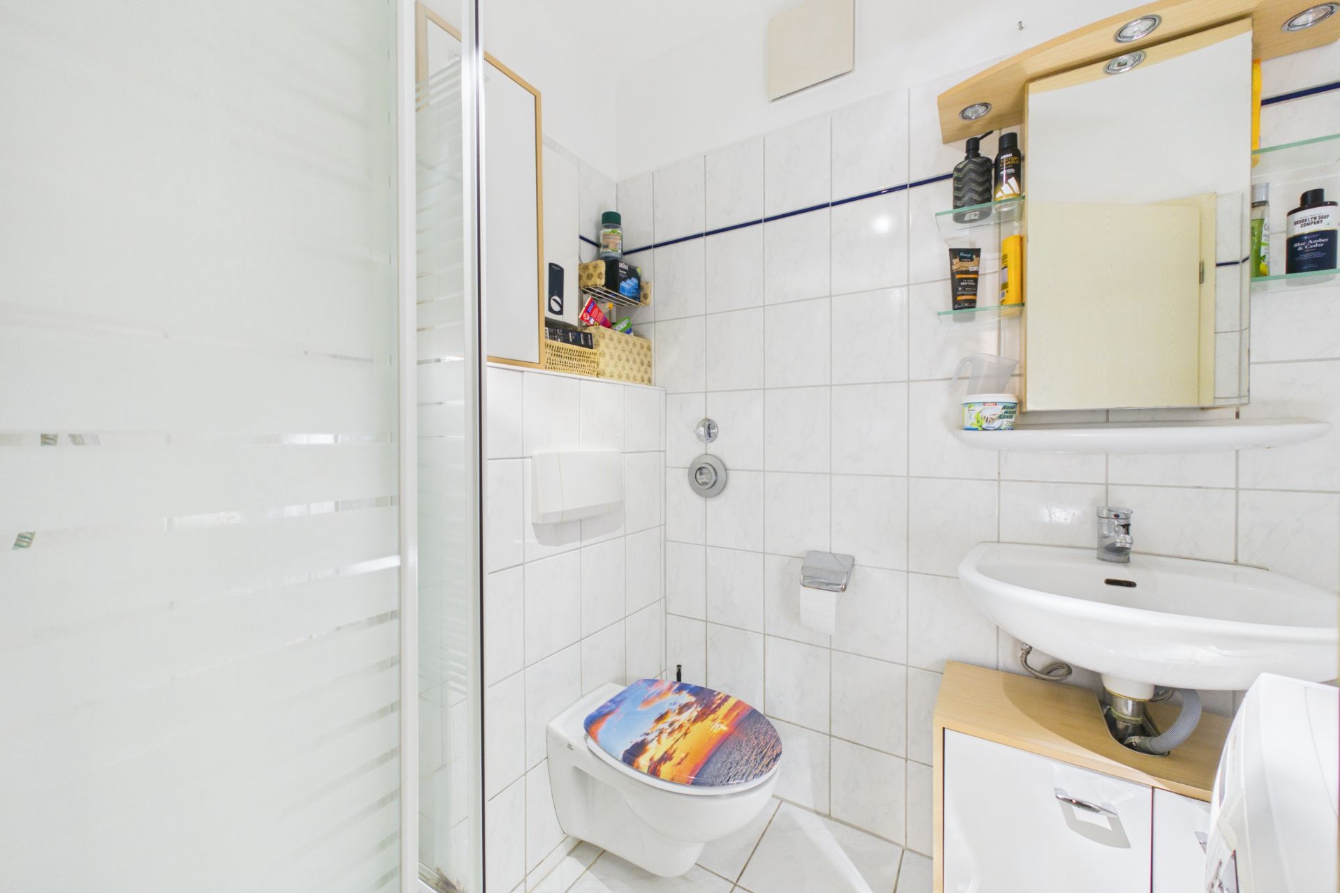 Helles Badezimmer mit Dusche, WC mit Motivdeckel, Waschbecken und Spiegelschrank.