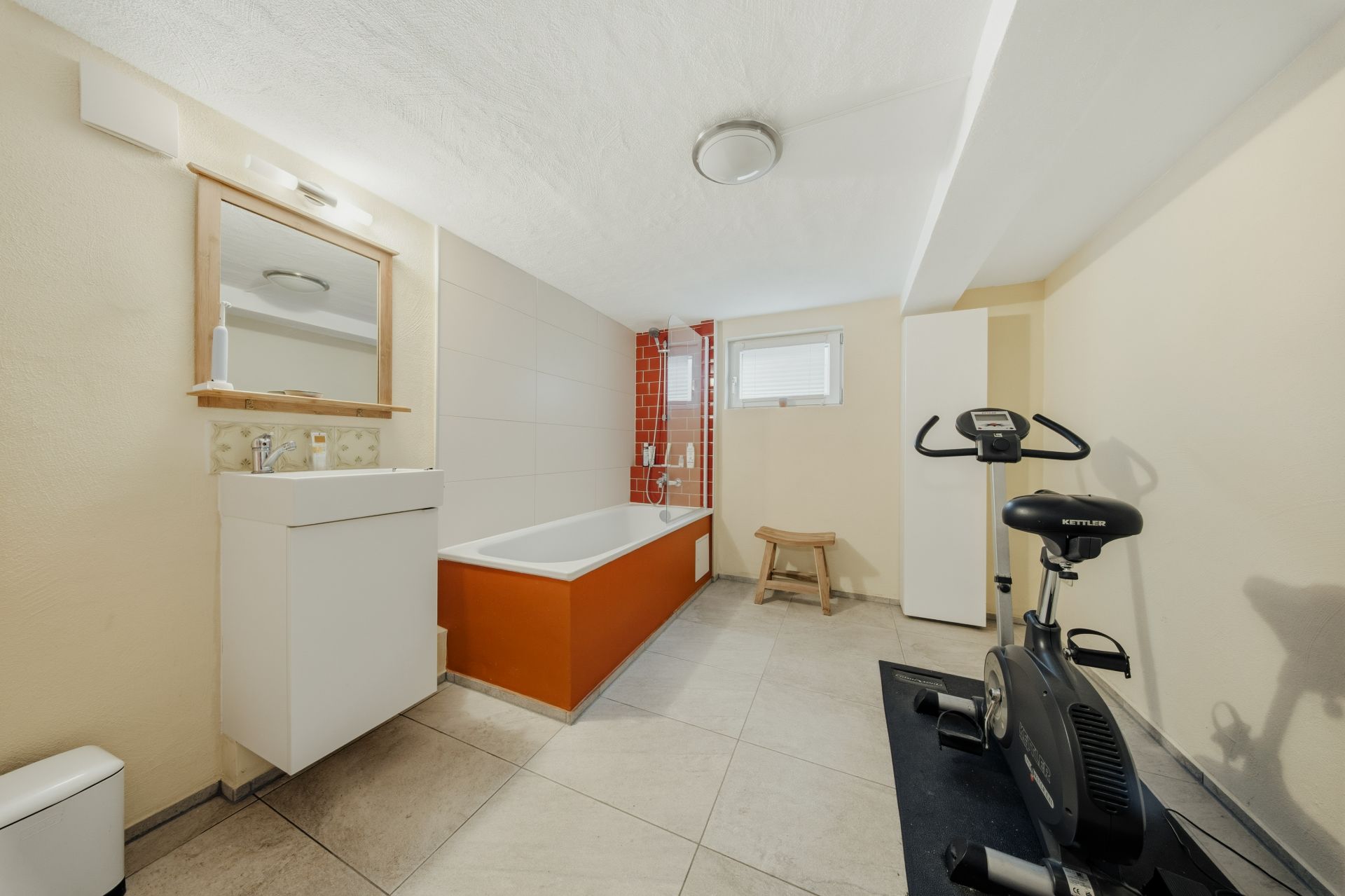 Badezimmer mit Badewanne, Waschbecken, Spiegelschrank und Heimtrainer auf Fliesenboden.