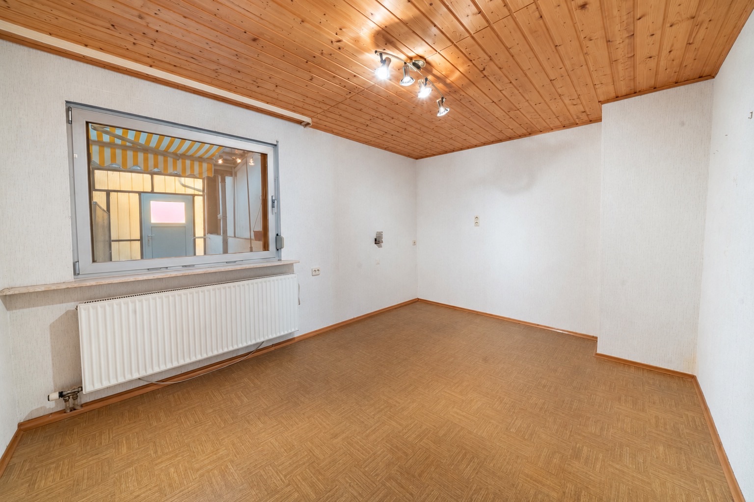 Leerer Raum mit Holzdecke, Fenster, Heizkörper und beigem Boden.