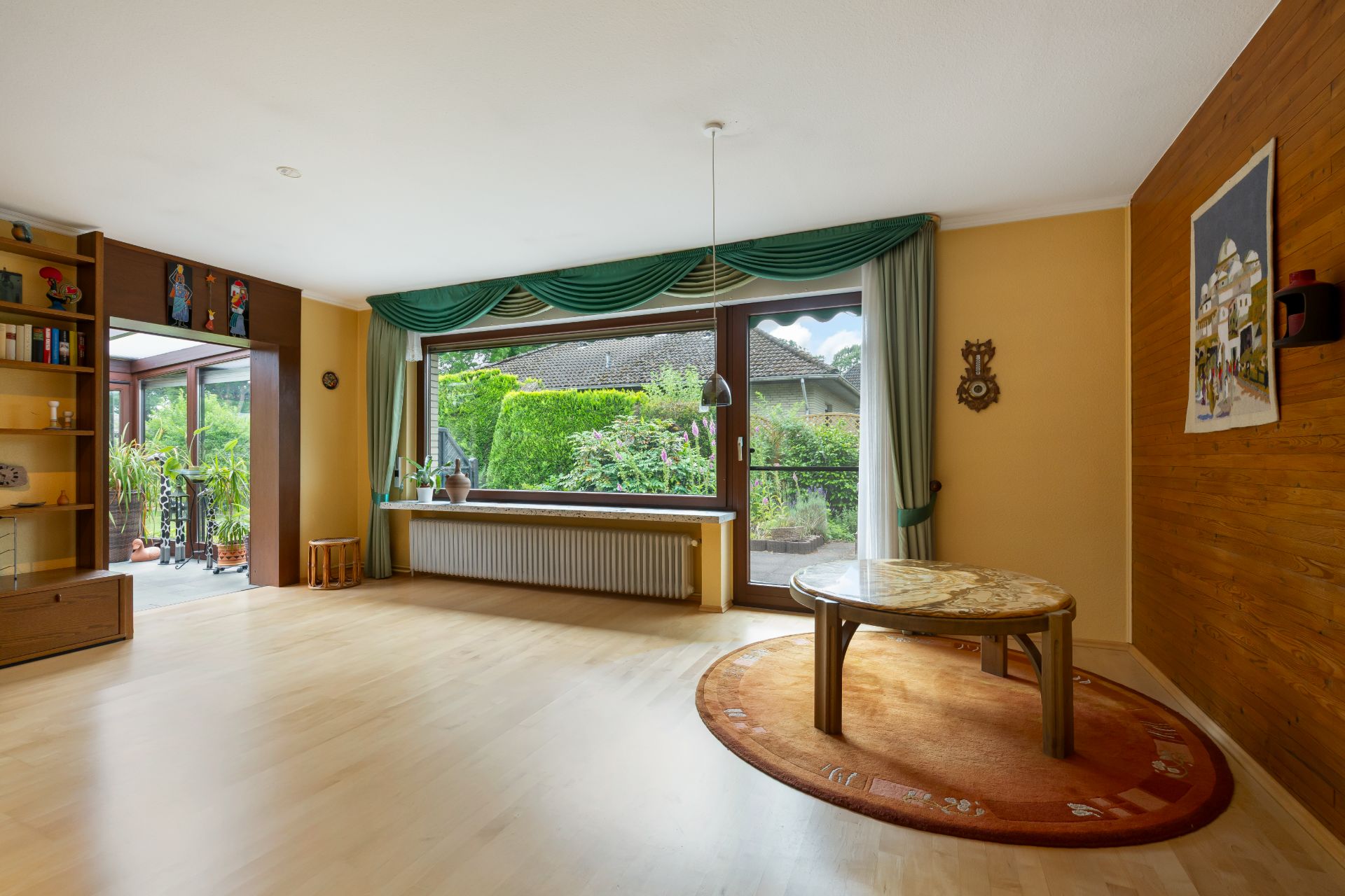 Helles Wohnzimmer mit großem Fenster, rundem Tisch auf Teppich und Regal mit Büchern.