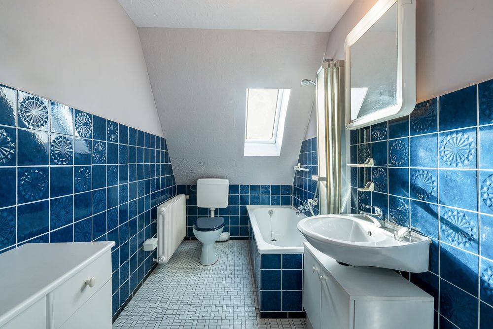 Badezimmer mit blauen Fliesen, Badewanne, Toilette und Waschbecken unter schrägem Dachfenster.
