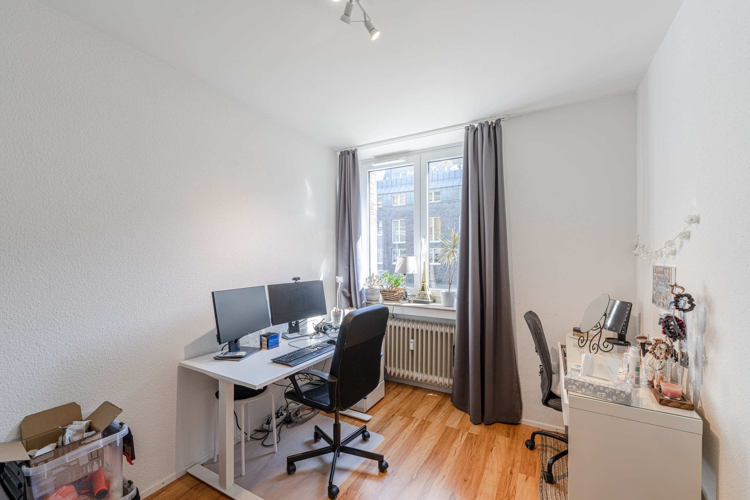 Helles Arbeitszimmer mit zwei Schreibtischen, Stühlen, Monitoren und großem Fenster mit grauen Vorhängen.