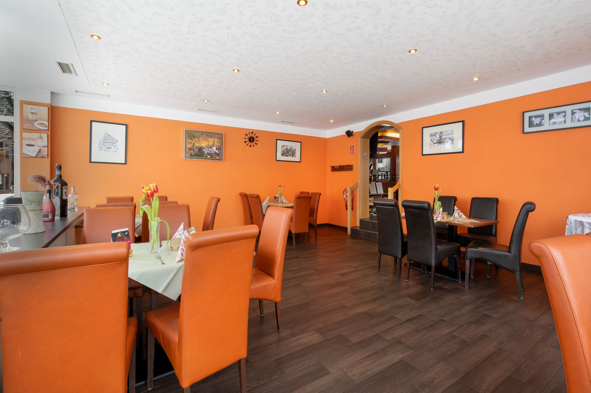 Esszimmer mit orangefarbenen Wänden, mehreren Tischen und Stühlen, Holzboden und Wandbildern.