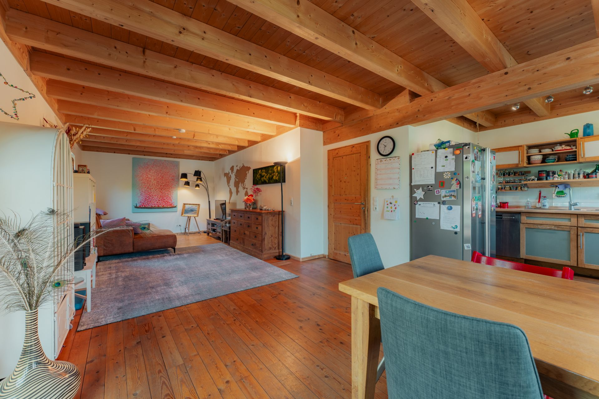 Offenes Wohnzimmer mit Holzboden, Esstisch, Sofa, Küche und sichtbaren Holzbalken an der Decke.