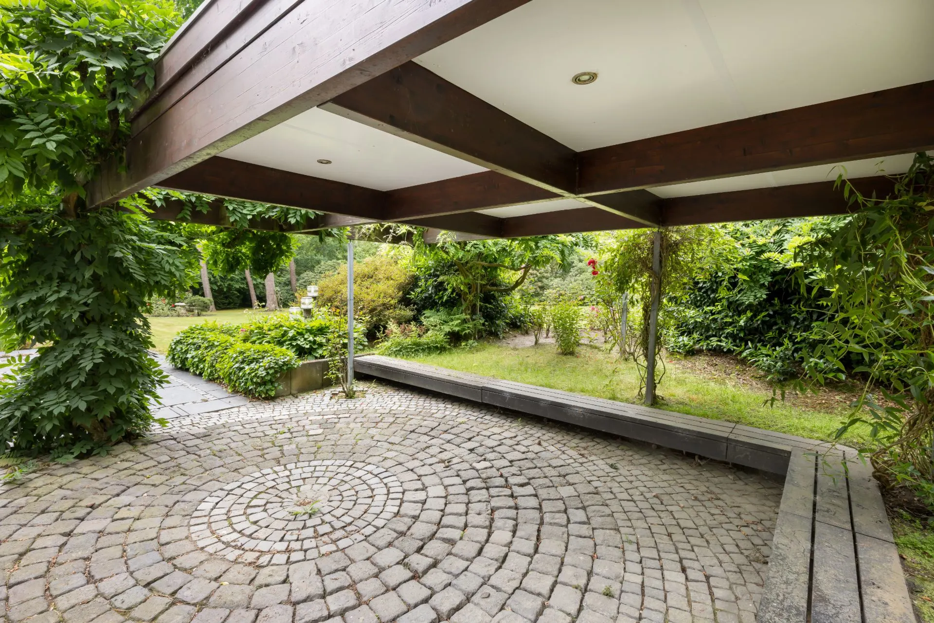 Überdachte Terrasse mit Pflastersteinen und Bank, umgeben von viel Grün und Pflanzen.