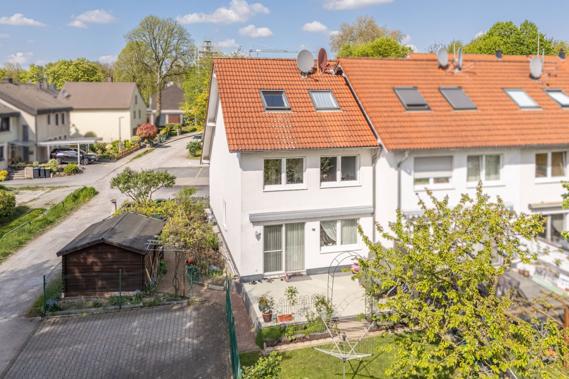 Reihenhaus mit rotem Dach, Garten und Terrasse, daneben ein Holzschuppen und Zufahrt.