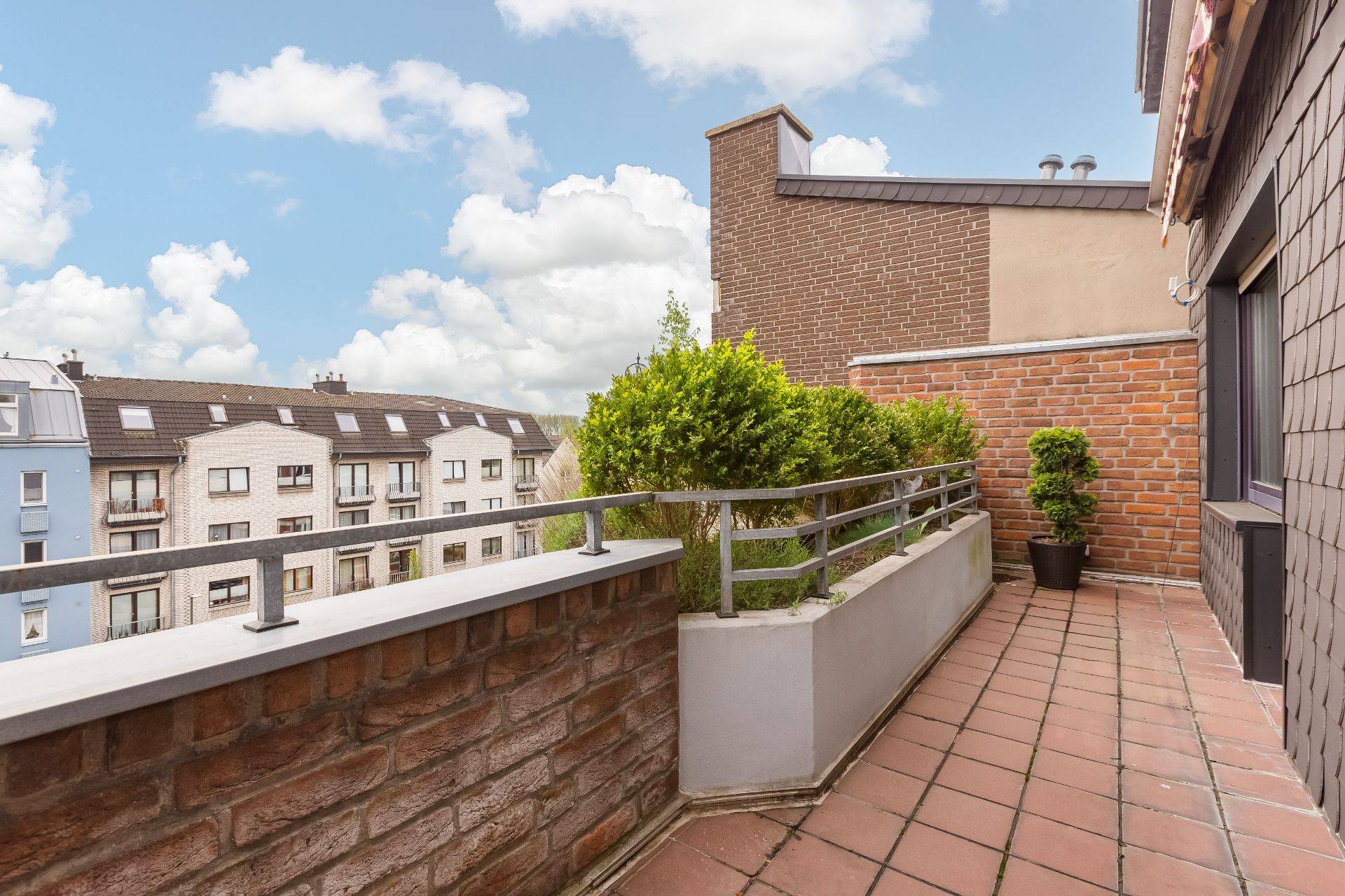 Balkon mit Pflanzkübeln, Ziegelwand und Blick auf benachbarte Wohnhäuser.