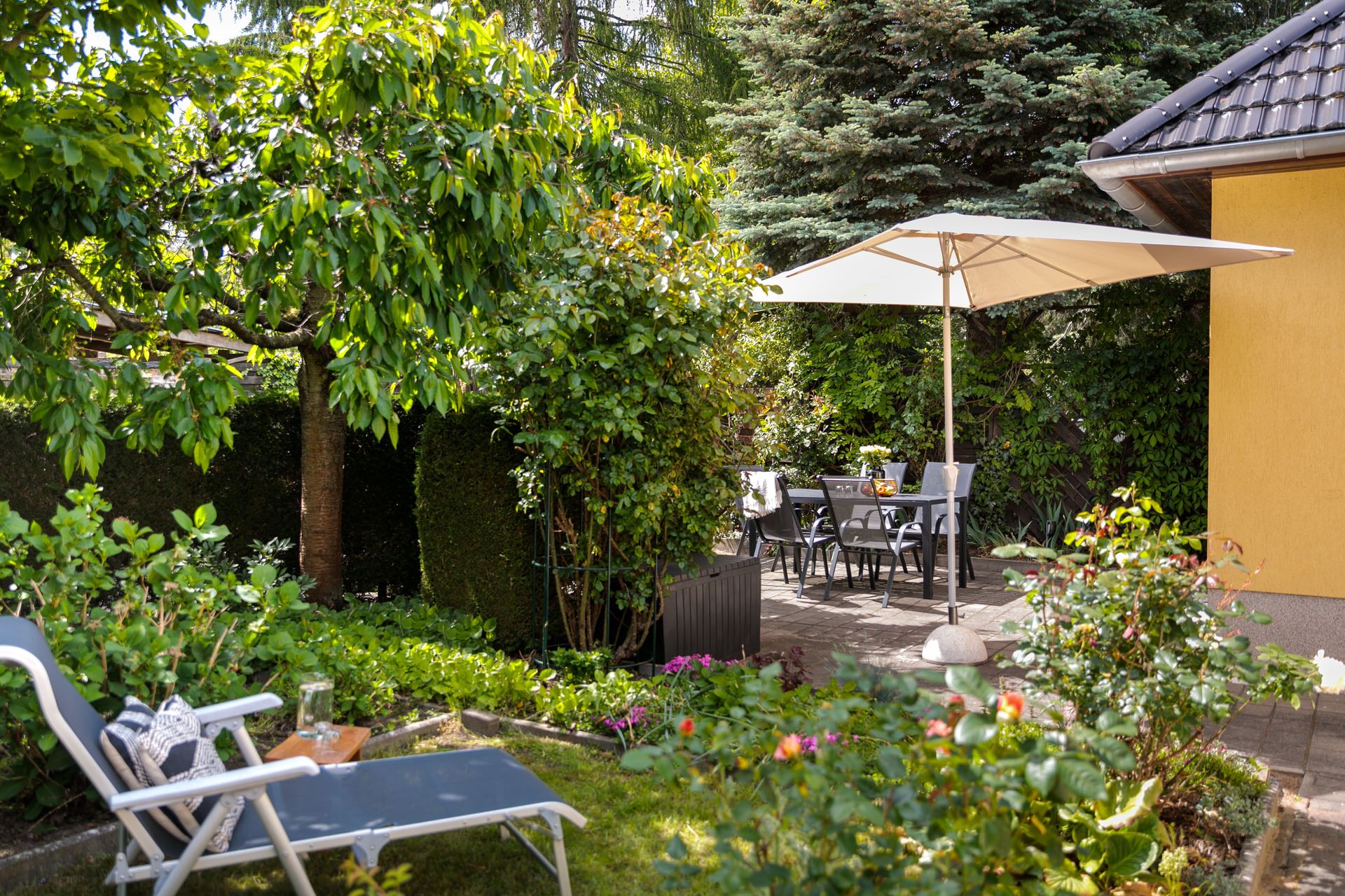 Garten mit Liegestuhl, Blumenbeeten und Sitzbereich unter einem Sonnenschirm neben einem gelben Haus.