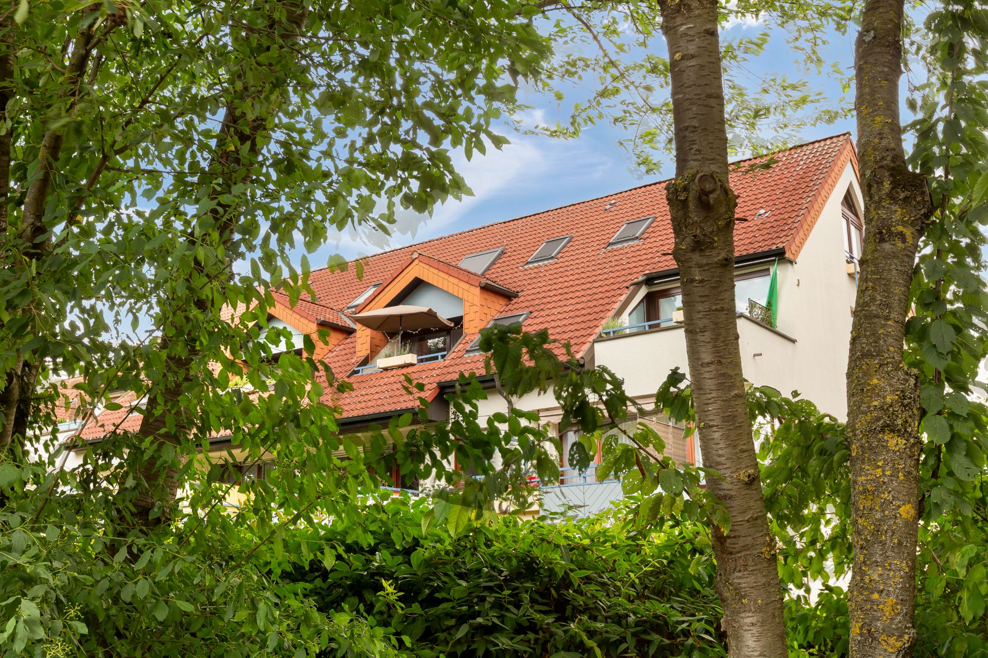 Wohnhaus mit rotem Dach hinter grünen Bäumen und Sträuchern.