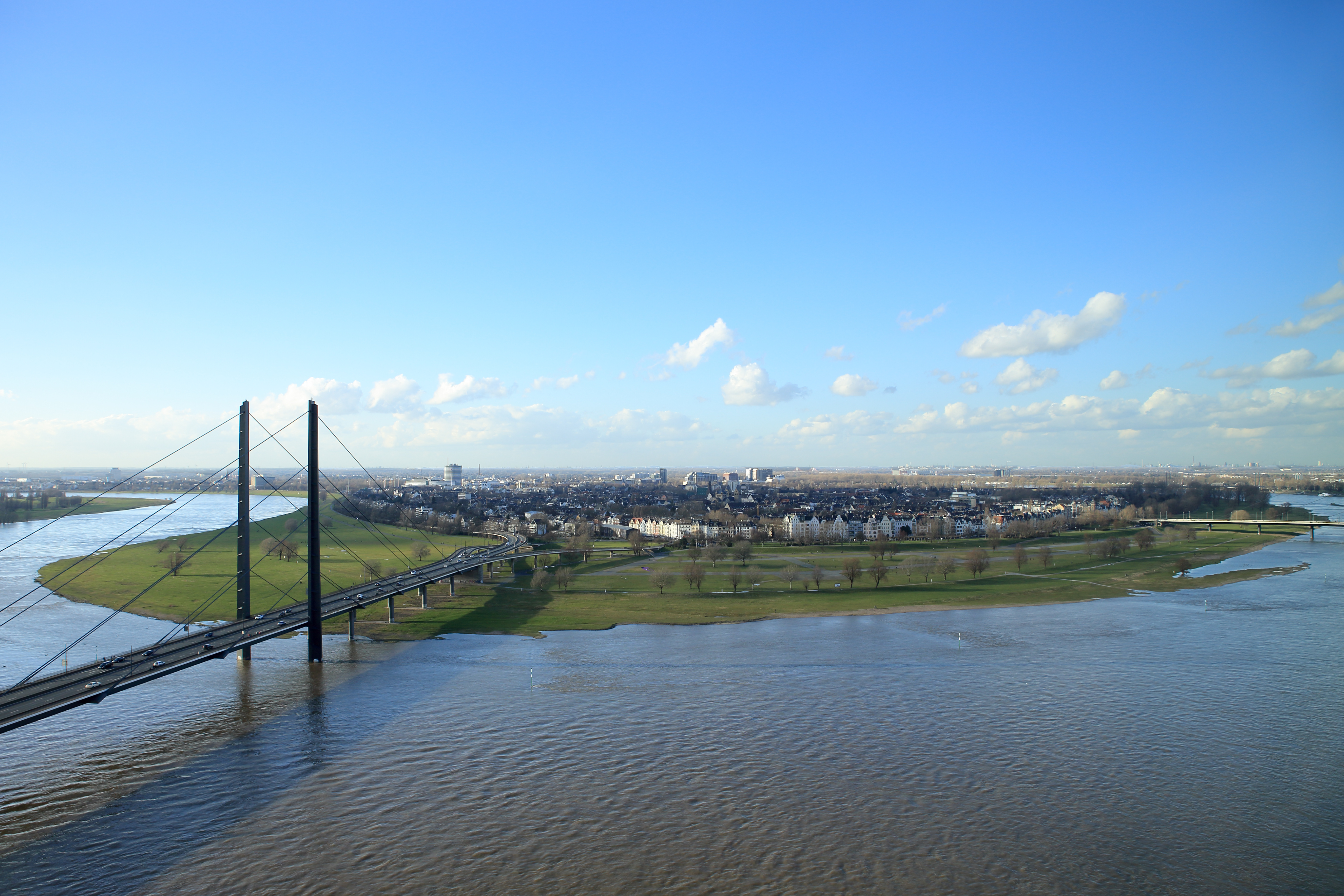 Aussicht auf eine Brücke über einen breiten Fluss, Grünflächen und eine Stadt im Hintergrund.