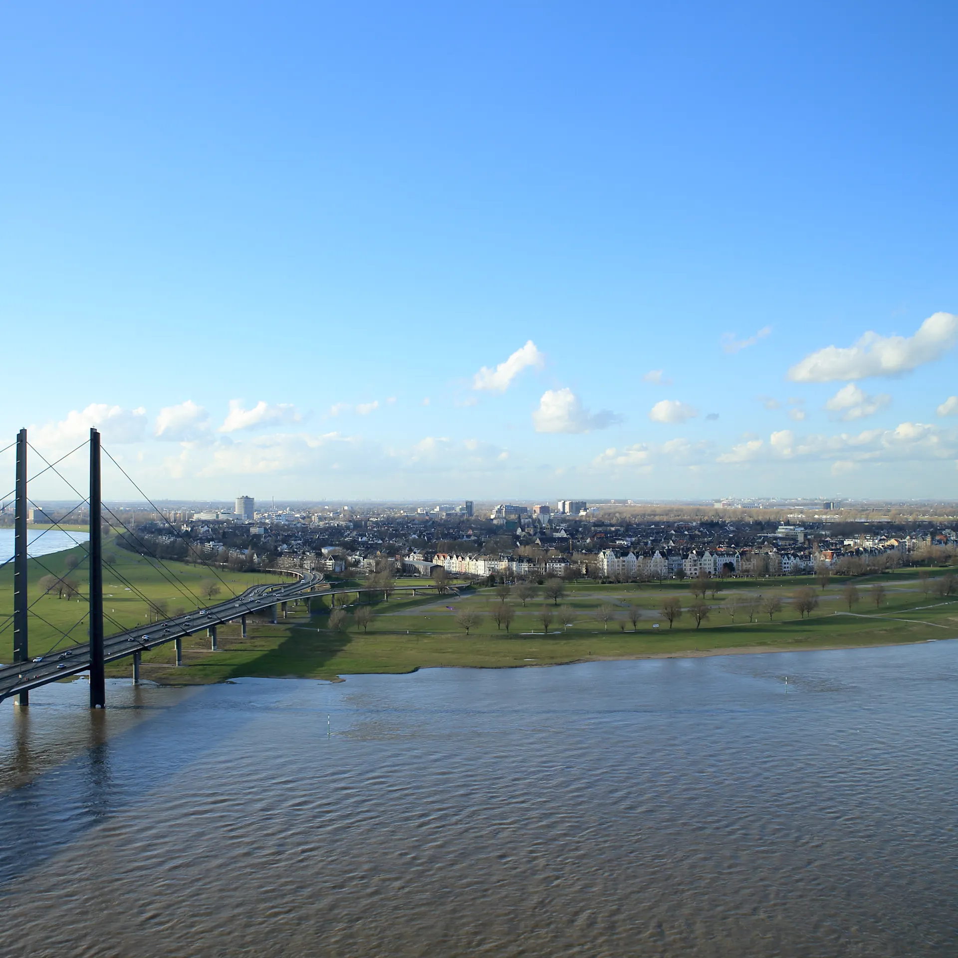 Aussicht auf eine Brücke über einen breiten Fluss, Grünflächen und eine Stadt im Hintergrund.