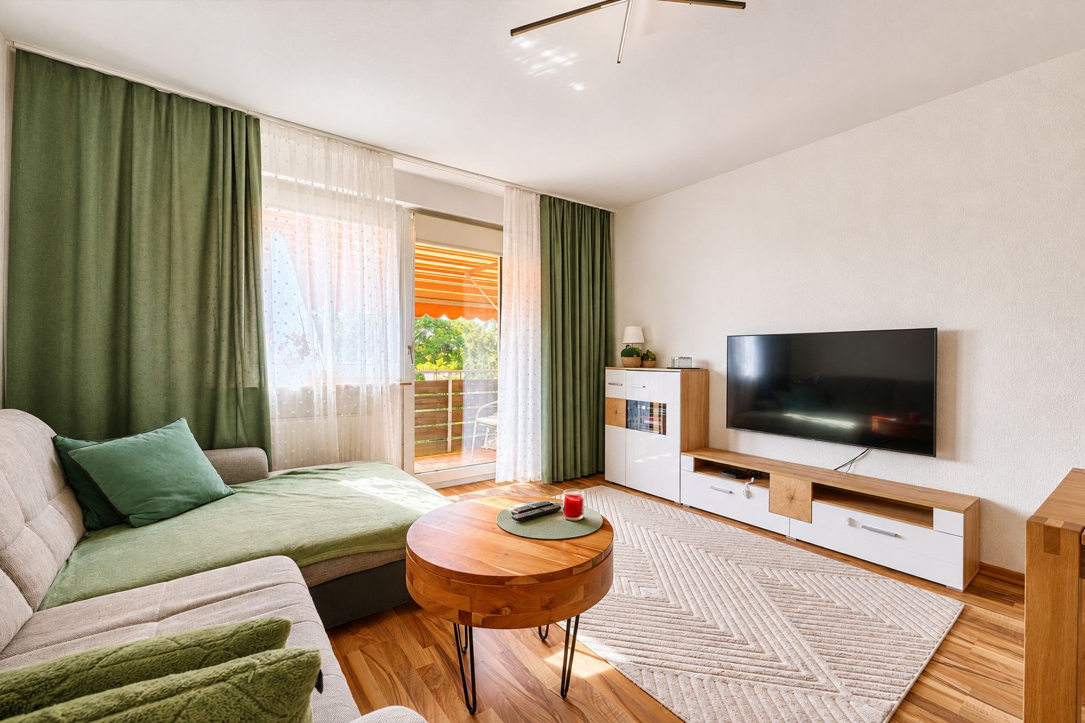 Helles Wohnzimmer mit Ecksofa, Couchtisch, Teppich und Fernseher, Zugang zum Balkon mit Vorhängen.