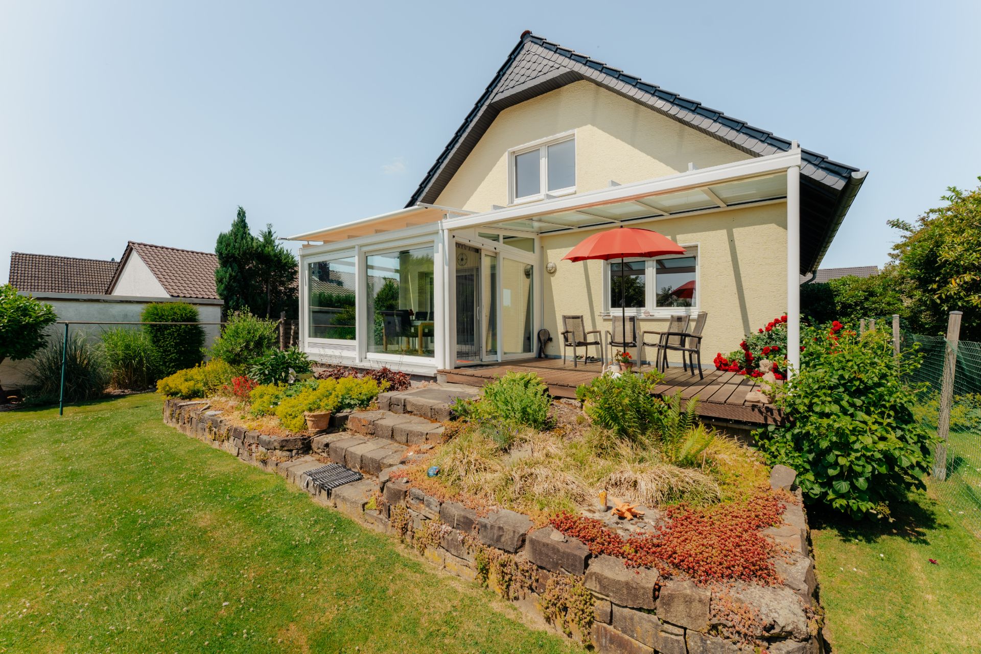 Einfamilienhaus mit Wintergarten, Terrasse, rotem Sonnenschirm und kleinem Garten mit Steinmauer.