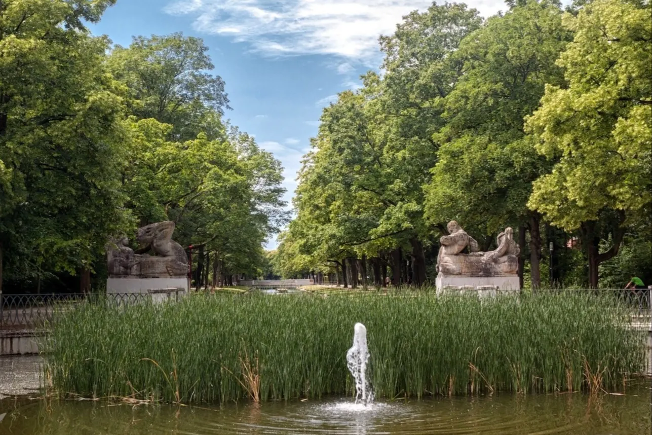 Springbrunnen vor hohen Büschen, flankiert von zwei Steinskulpturen, umgeben von vielen Bäumen.