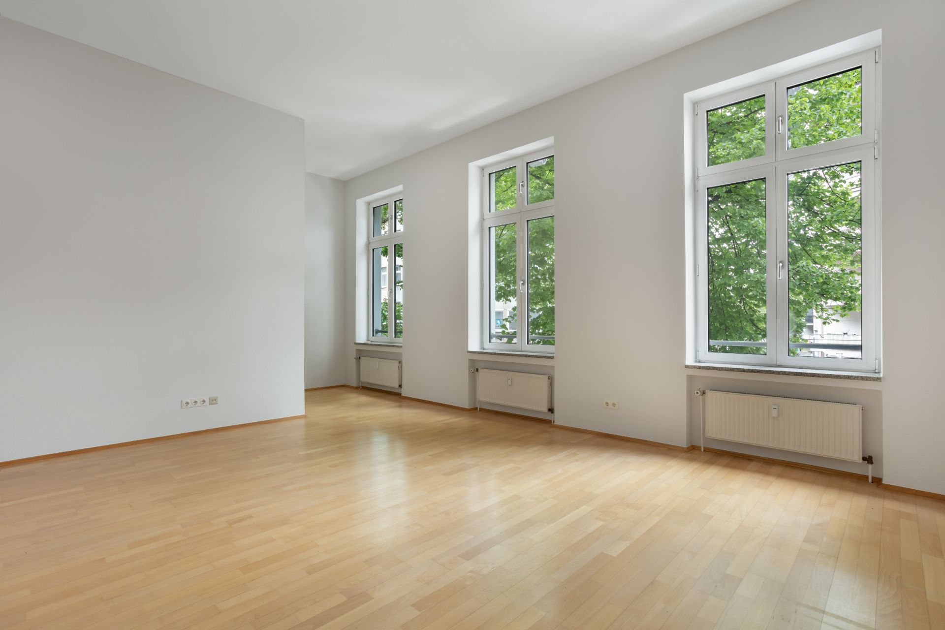 Heller, leerer Raum mit Holzfußboden und drei großen Fenstern.