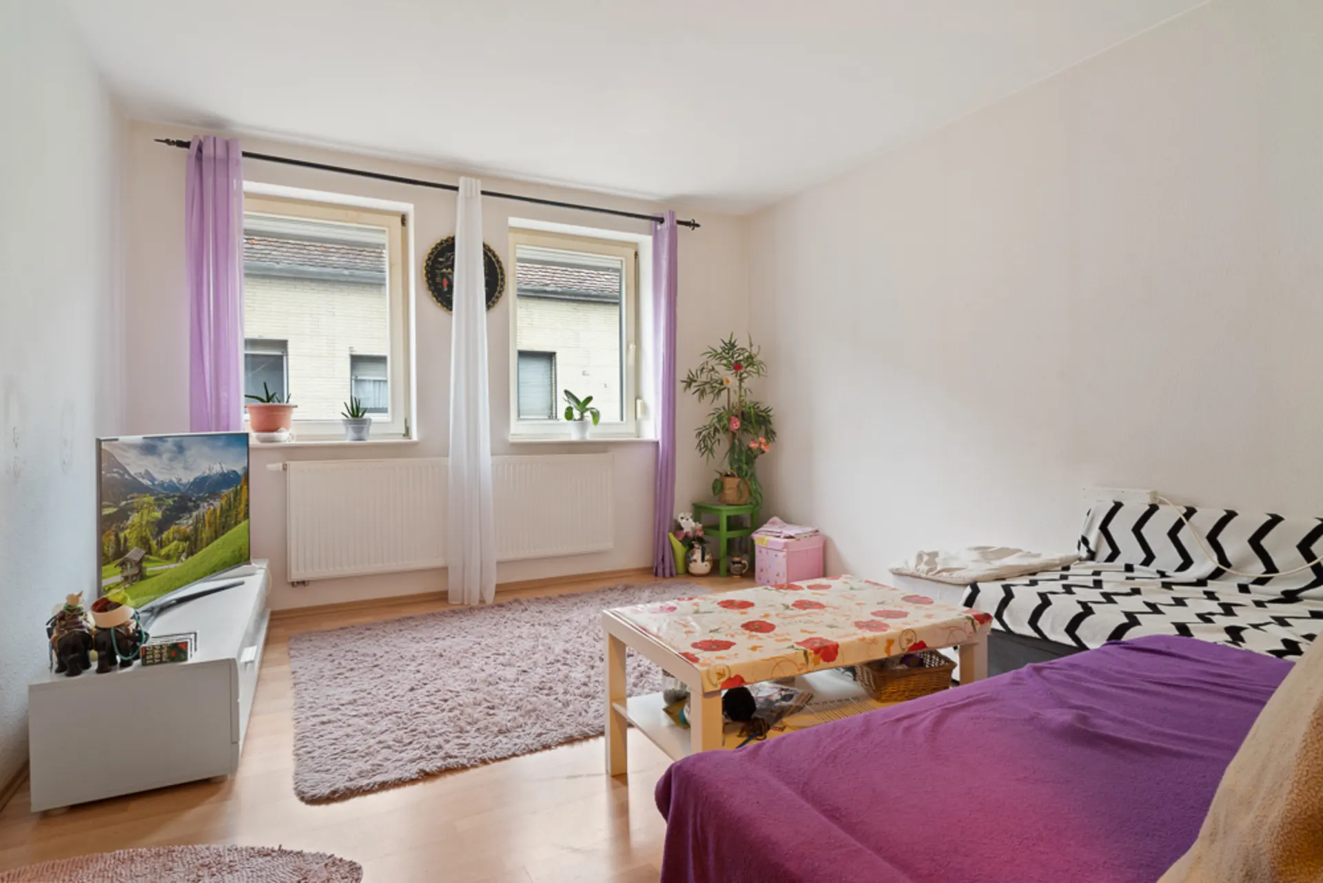 Helles Wohnzimmer mit zwei Fenstern, Sofa, Couchtisch, Fernseher und Pflanzen.