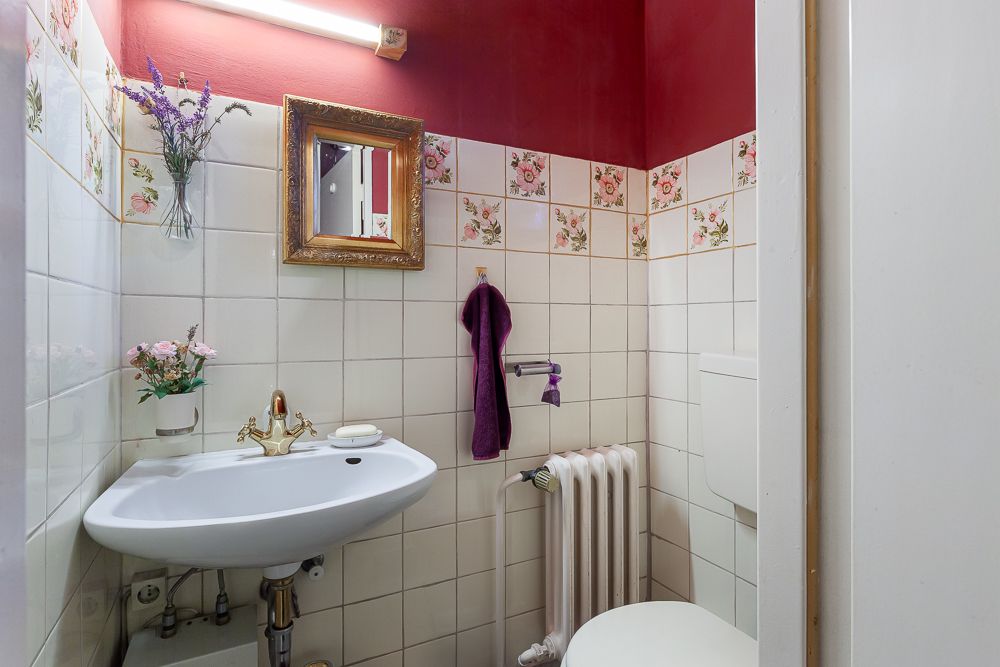 Kleines Badezimmer mit Blumenfliesen, Waschbecken, Spiegel, WC und Dekoblumen.