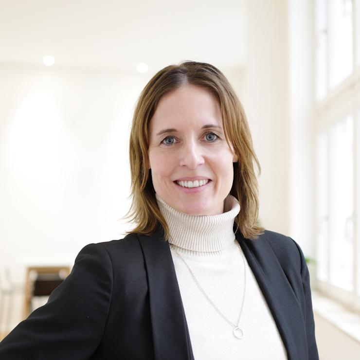 Frau mit hellen Haaren steht in einem hellen Raum mit großen Fenstern.