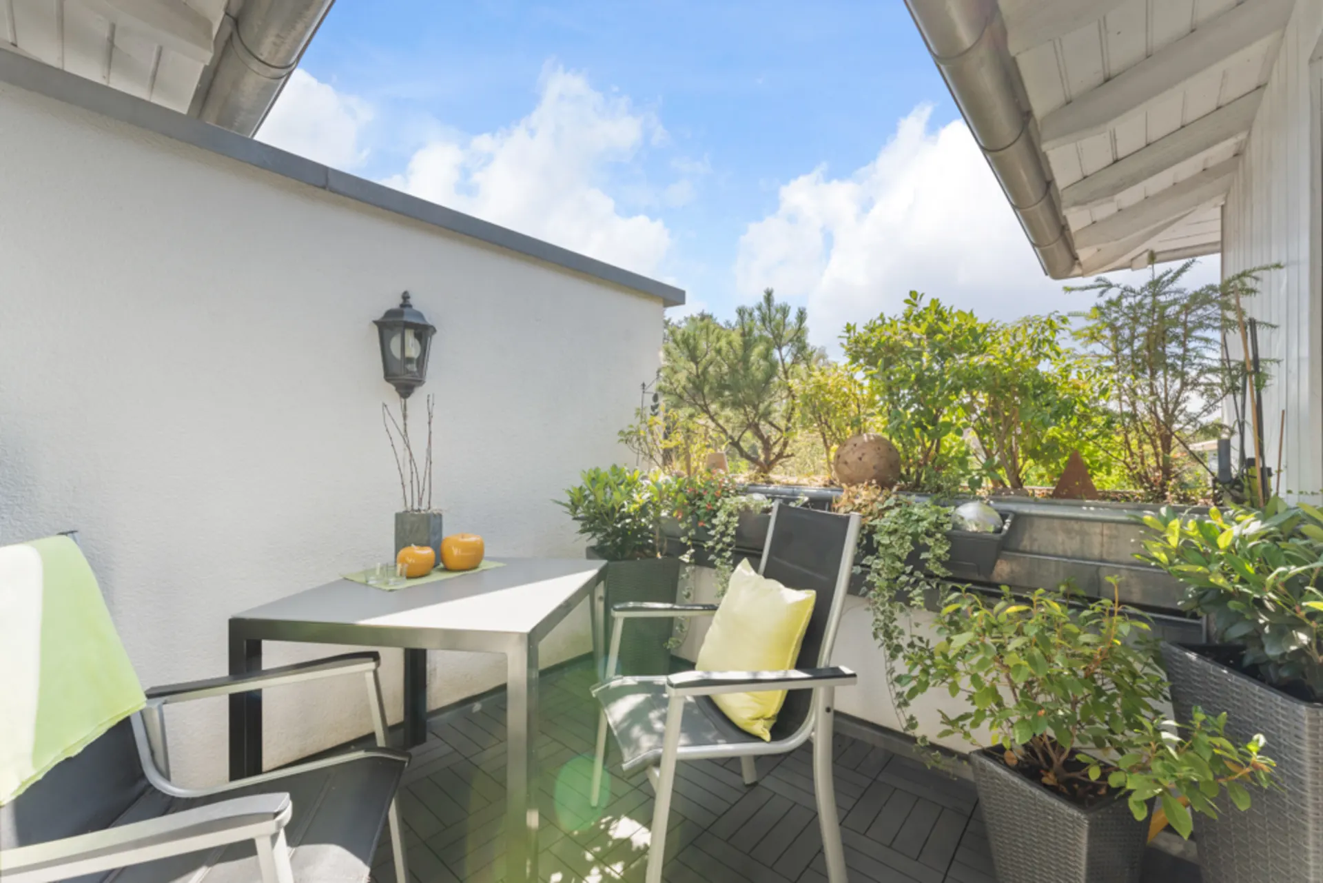 Kleine Terrasse mit zwei Stühlen, Tisch und vielen Pflanzen unter blauem Himmel.