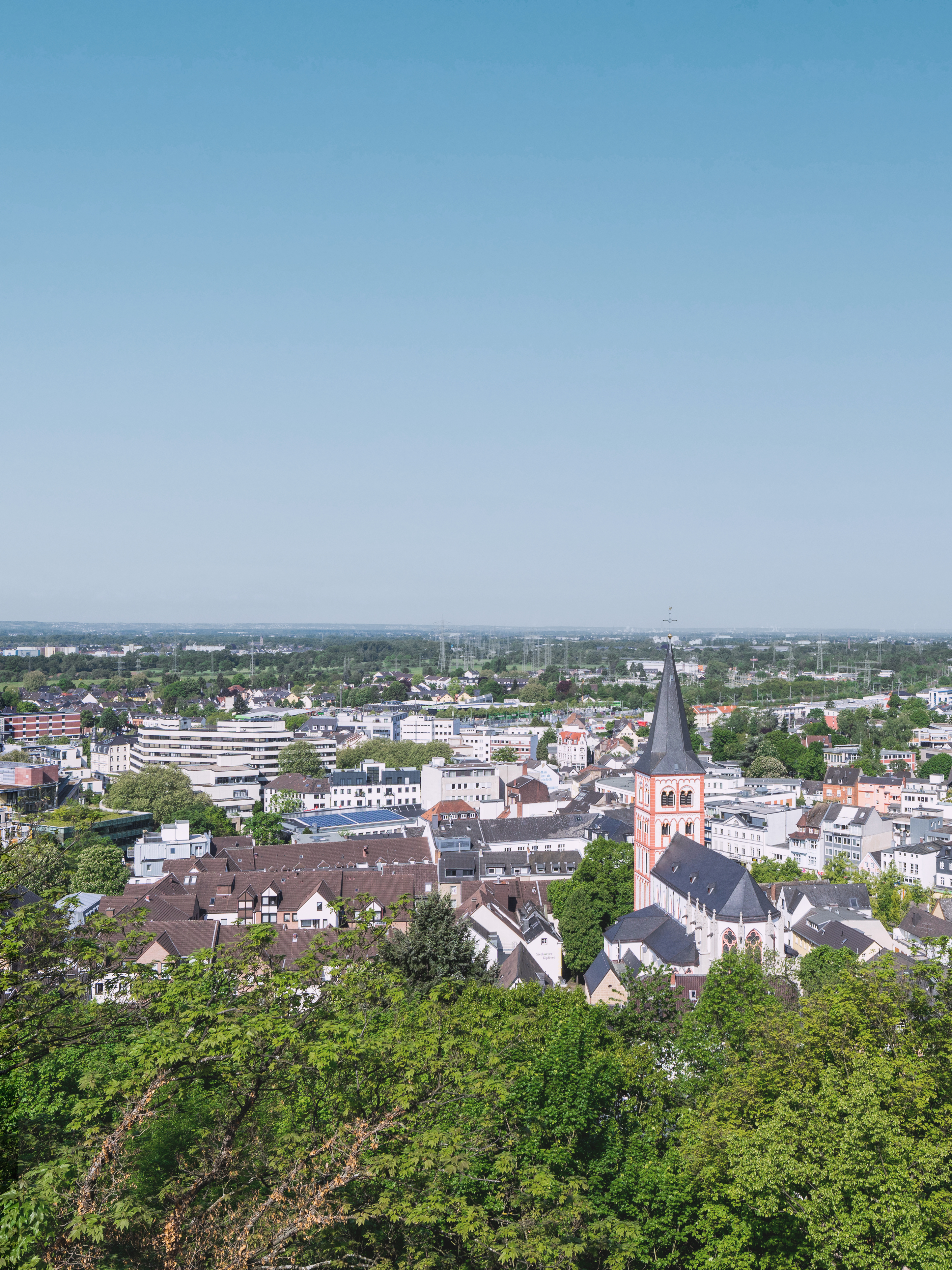 Stadtansicht mit vielen Häusern, einer Kirche mit Turm und viel grünem Baumbestand im Vordergrund.
