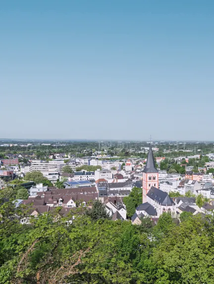 Stadtansicht mit vielen Häusern, einer Kirche mit Turm und viel grünem Baumbestand im Vordergrund.